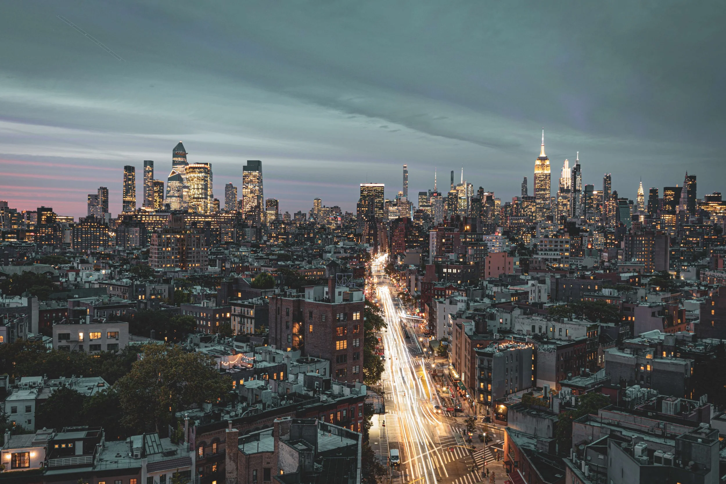 Nighttime view of New York City skyline with illuminated skyscrapers and traffic lights on a busy street.