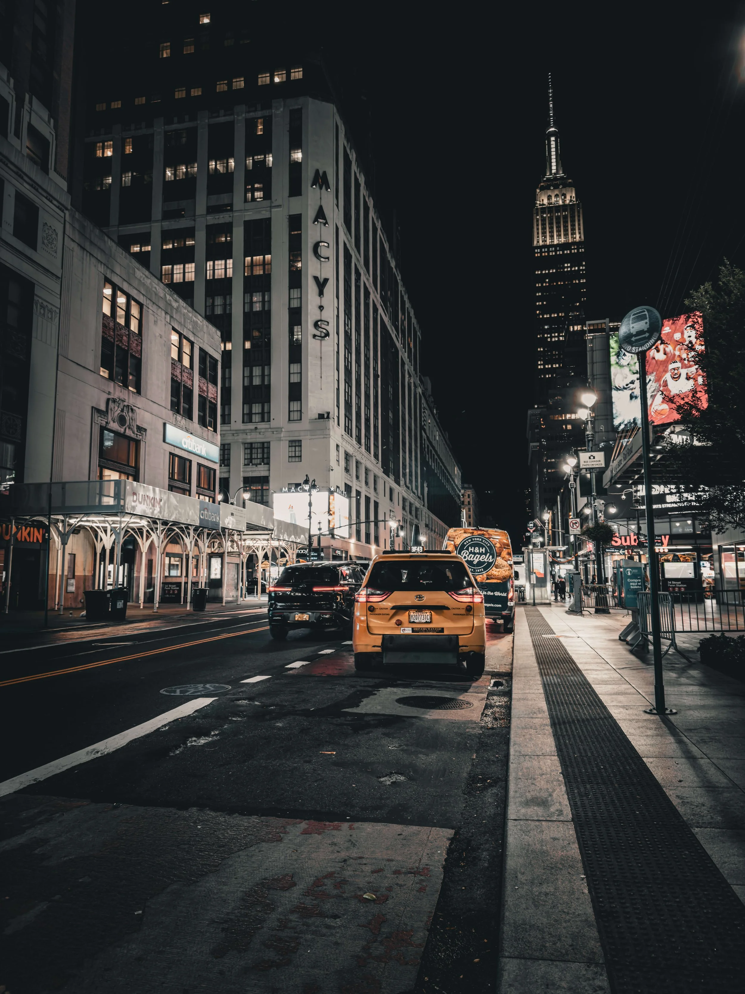 Nighttime city street with parked cars, illuminated buildings, a prominent sign reading 'MACY'S,' and the Empire State Building in the background.
