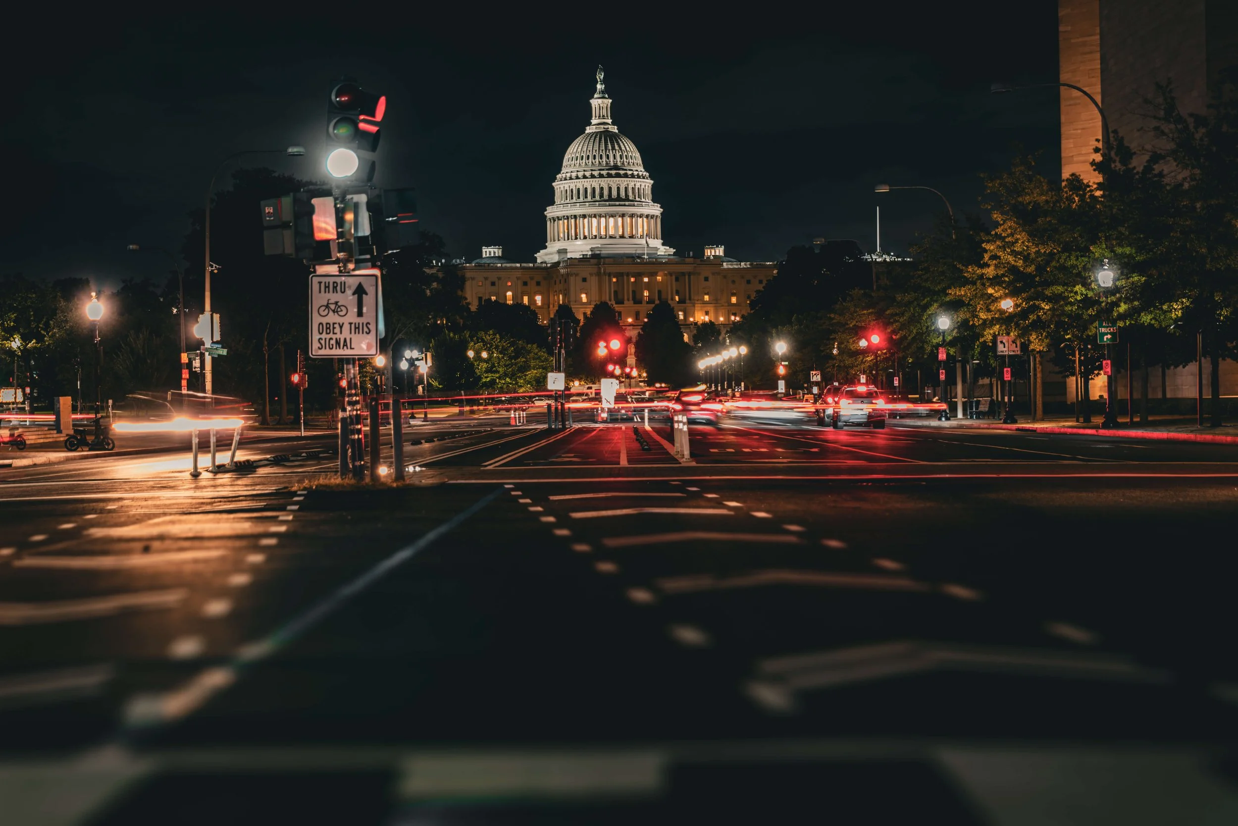 Nighttime view of the U.S. Capitol building in Washington, D.C., seen from a busy street with cars and traffic lights, illuminated by streetlights.