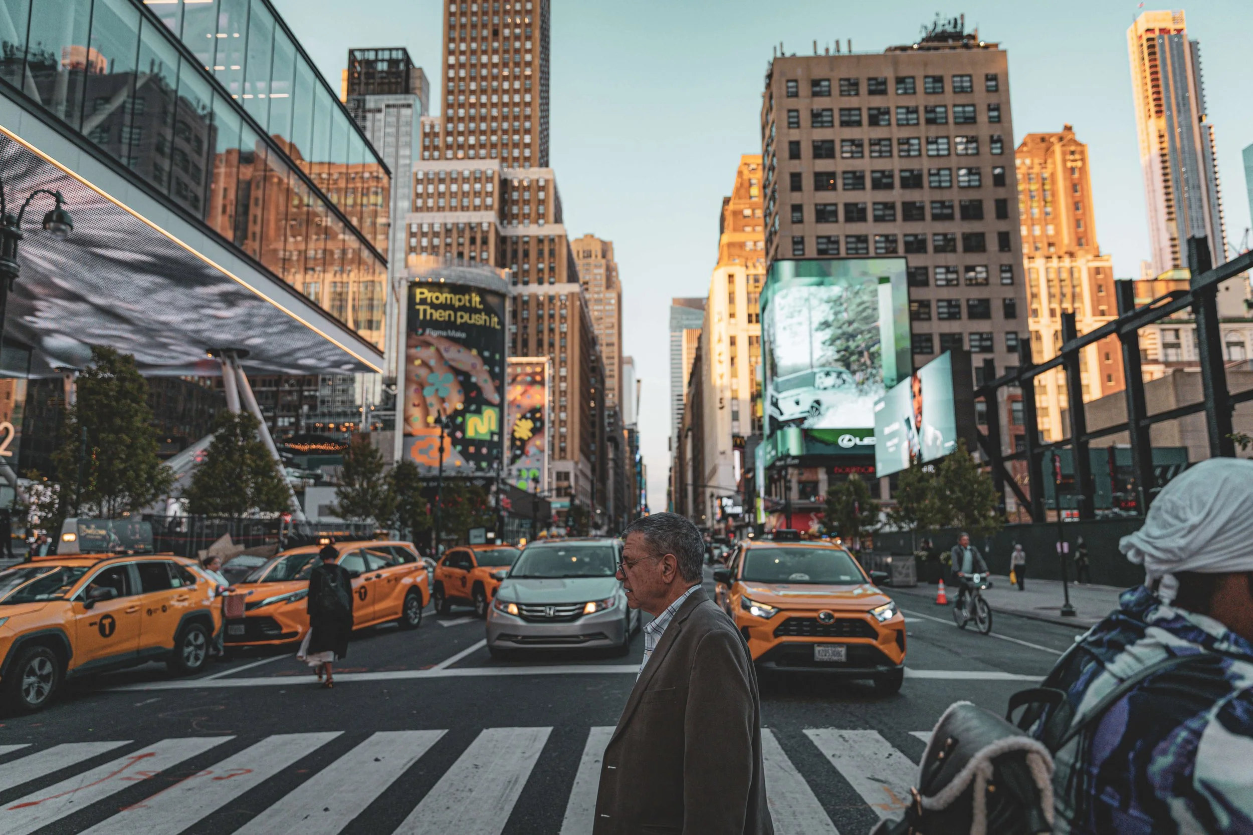 A busy city street with yellow taxis, pedestrians crossing, and tall buildings with billboards in the evening light.