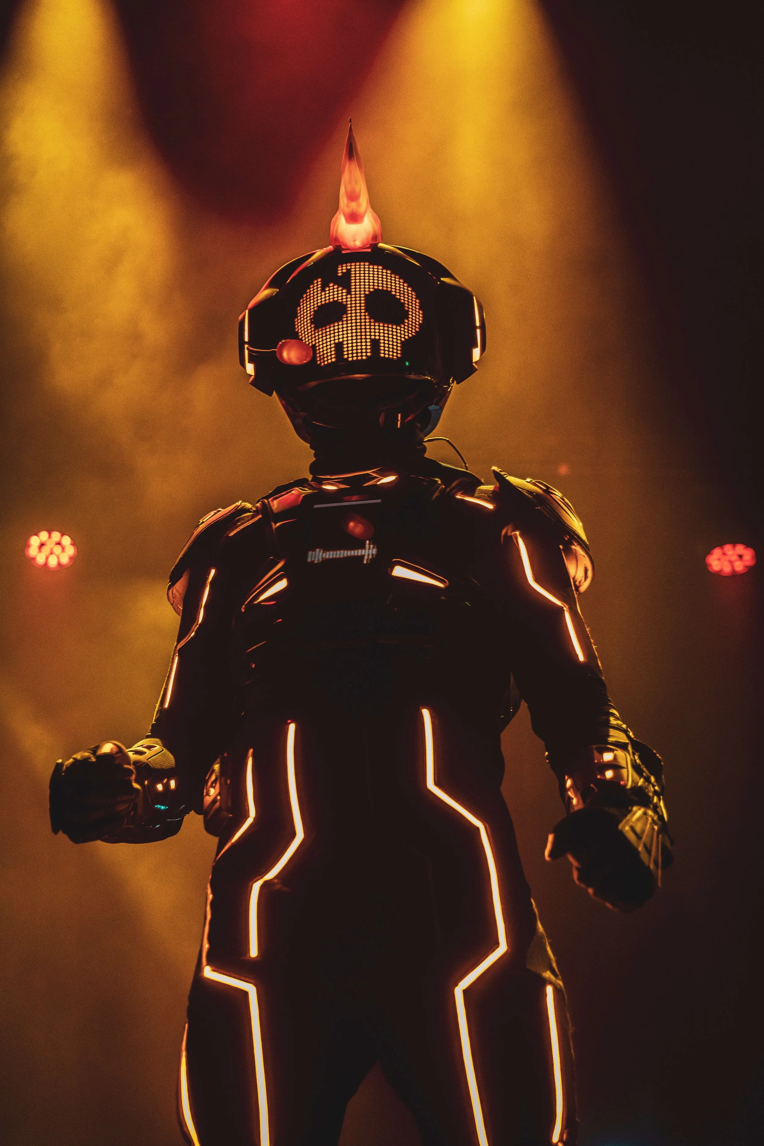 Person in a glowing black suit with neon lights and a helmet featuring a skull pattern, standing in a dimly lit environment with smoke or fog.