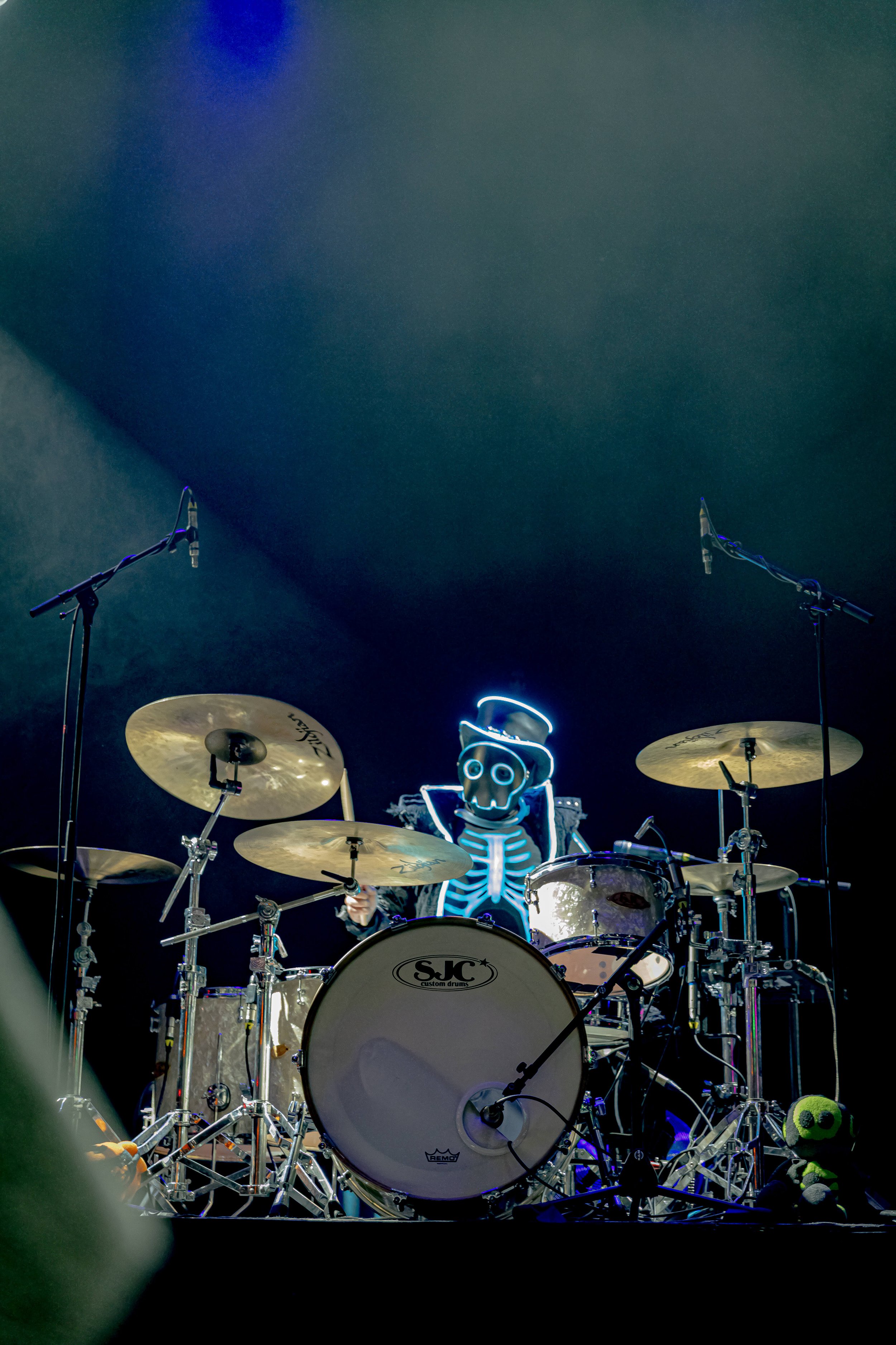 A drum set on stage with a person wearing a skeleton costume with neon lights and a skull mask behind  the drums. There are microphones and a stuffed frog toy nearby.