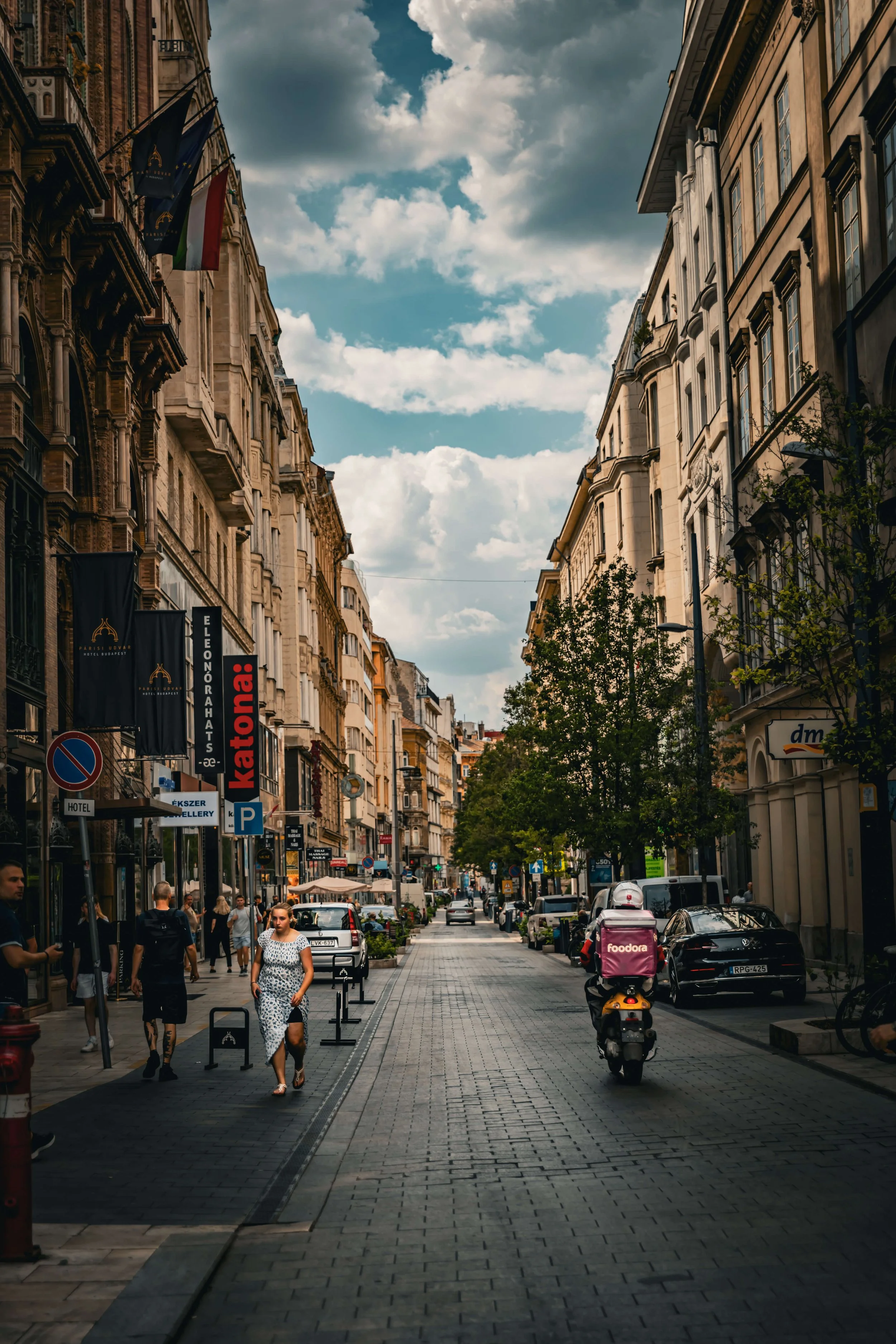 City street with shops, pedestrians, parked cars, a scooter, and a partly cloudy sky.