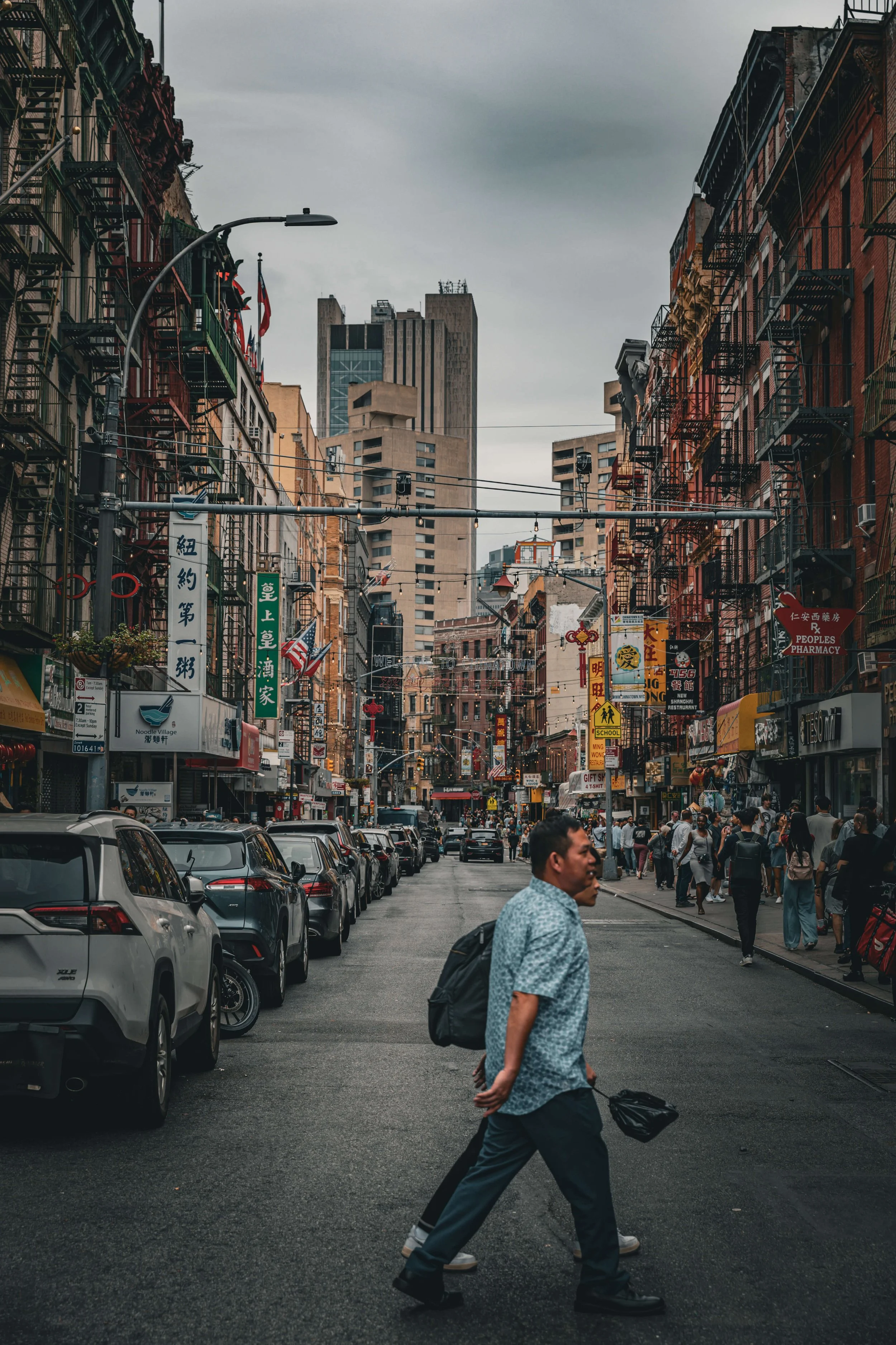 A busy city street with parked cars along the curb and people walking on sidewalks. Tall buildings line both sides, decorated with colorful signs and banners, with a mix of modern and older architectural styles. The sky is overcast.