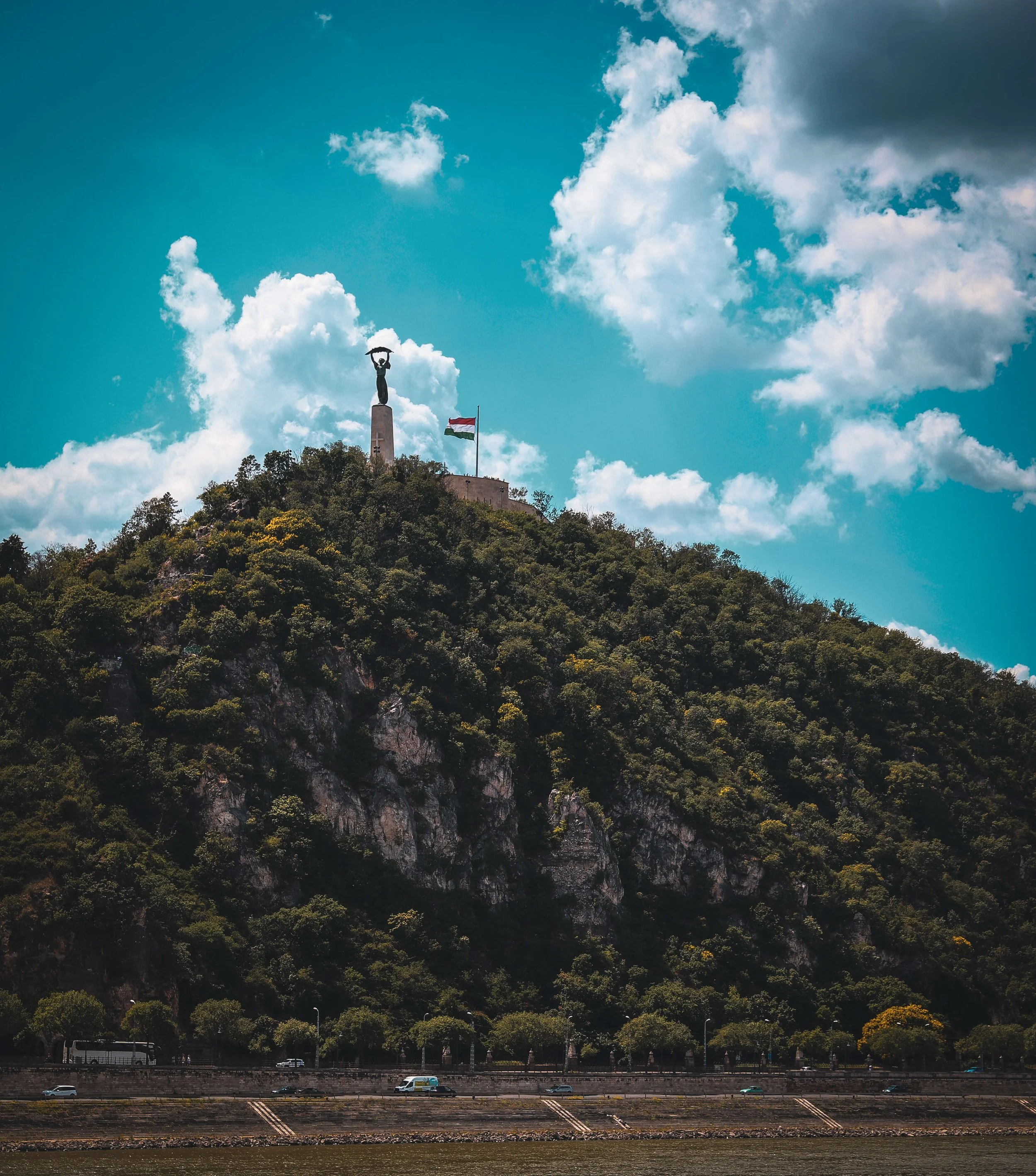 Statue of a woman holding a hat on a monument on a hill with green trees, a flag, and a partly cloudy blue sky.