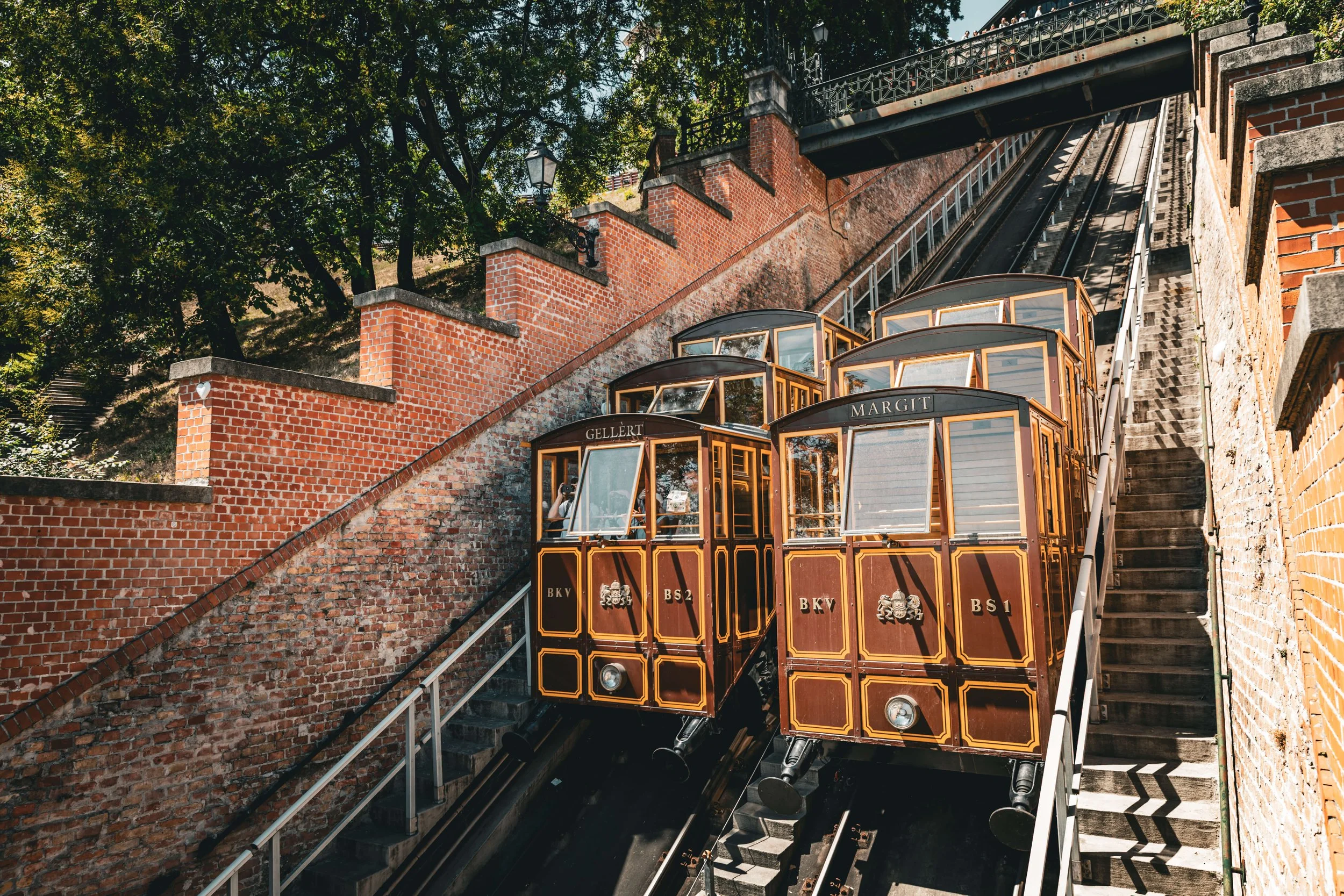 A funicular train ascending a brick hillside along a steep track with brick and stone walls, surrounded by trees, under a clear sky.