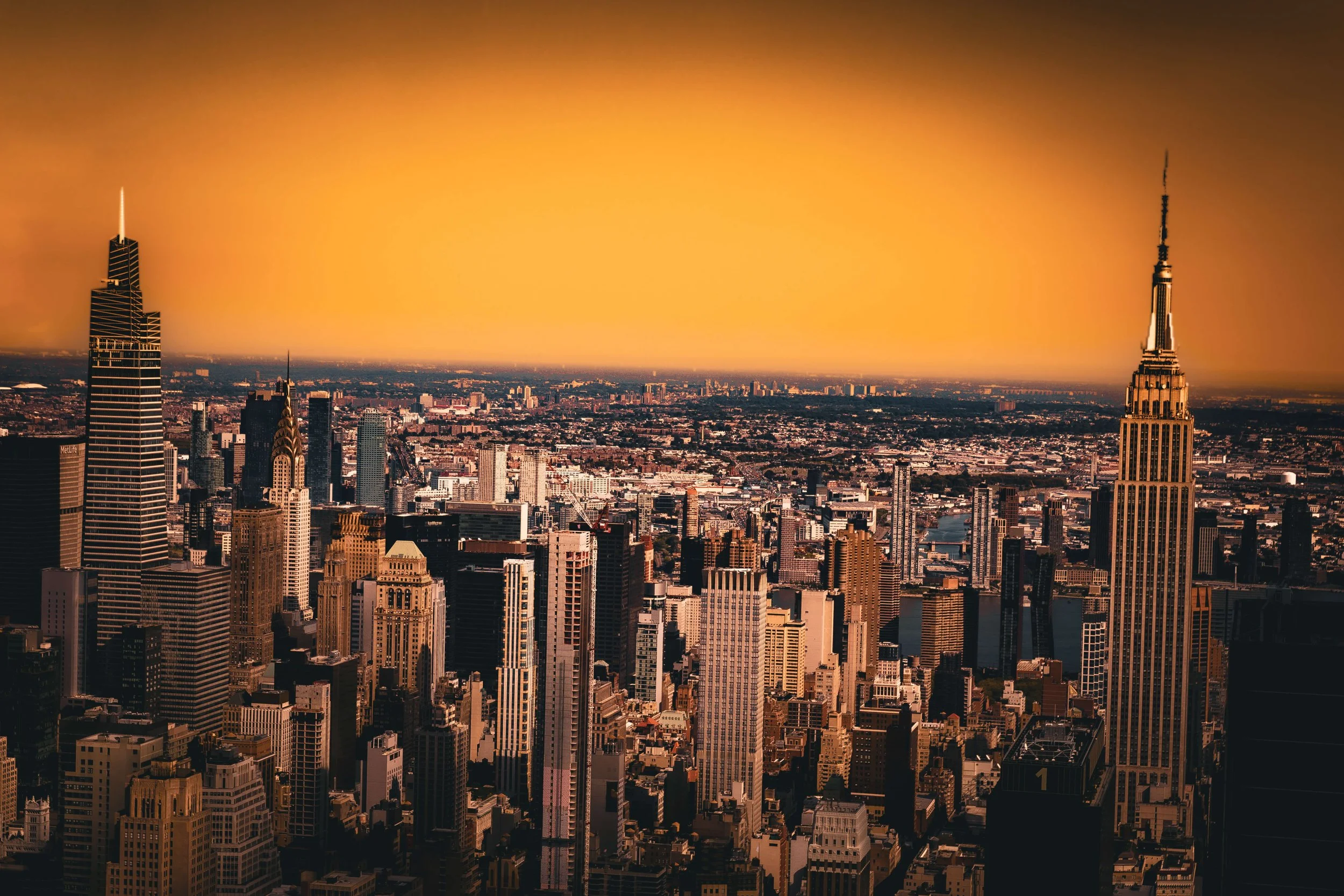 Aerial view of New York City skyline at sunset with tall skyscrapers, including Empire State Building, and a river visible in the background.