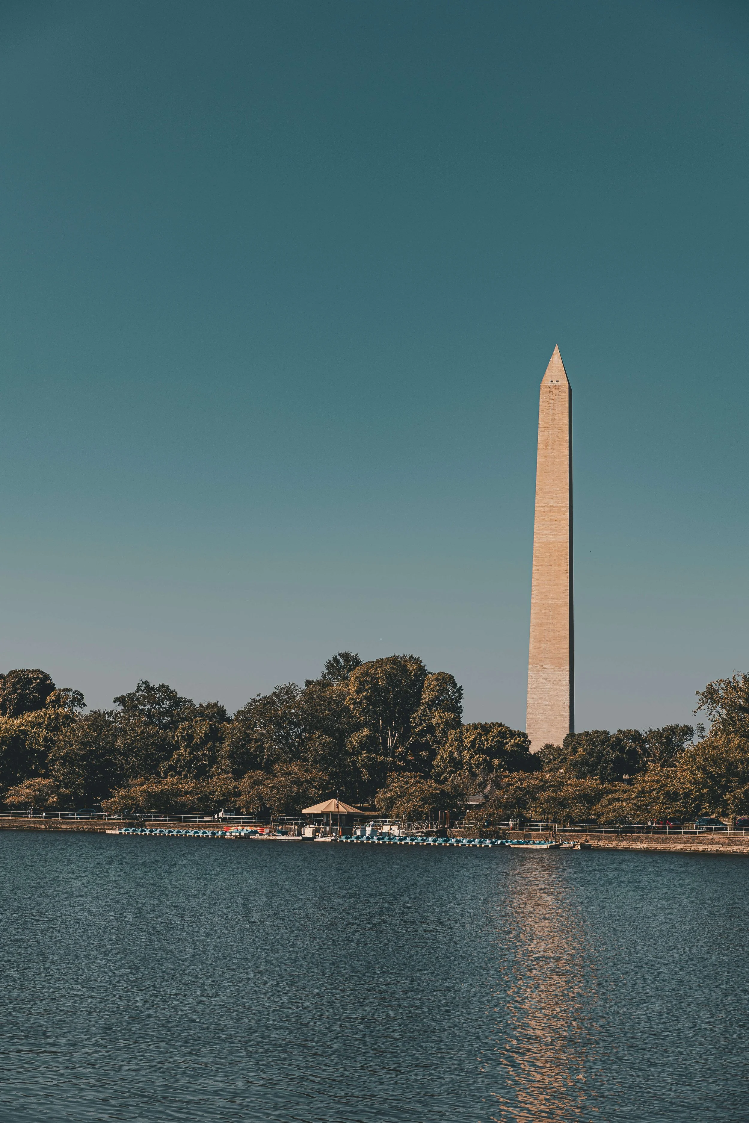 Washington Monument with a body of water in the foreground and trees surrounding it under a clear blue sky.