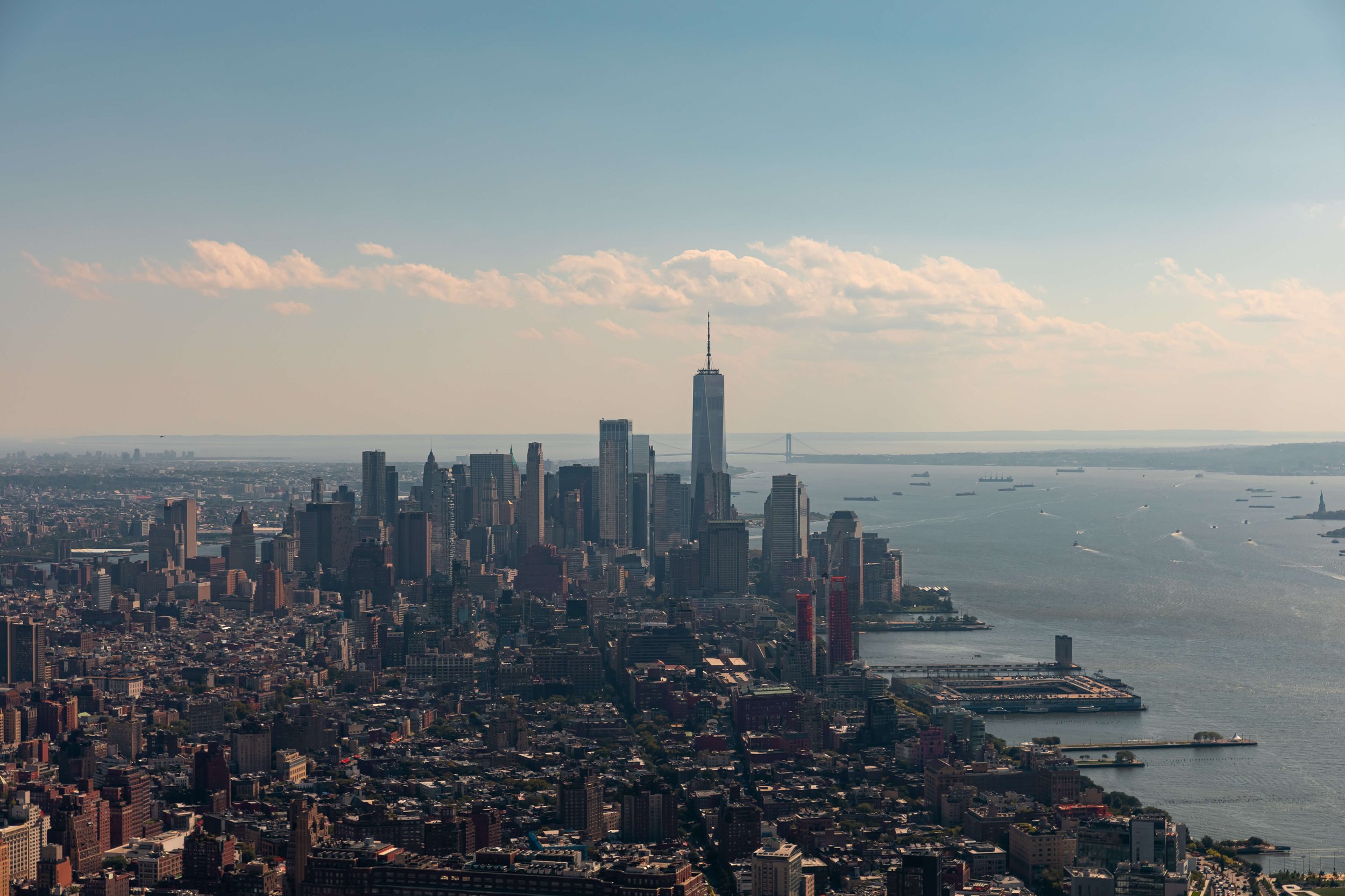 Aerial view of New York City skyline with One World Trade Center, skyscrapers, water, and bridges in the background on a partly cloudy day.