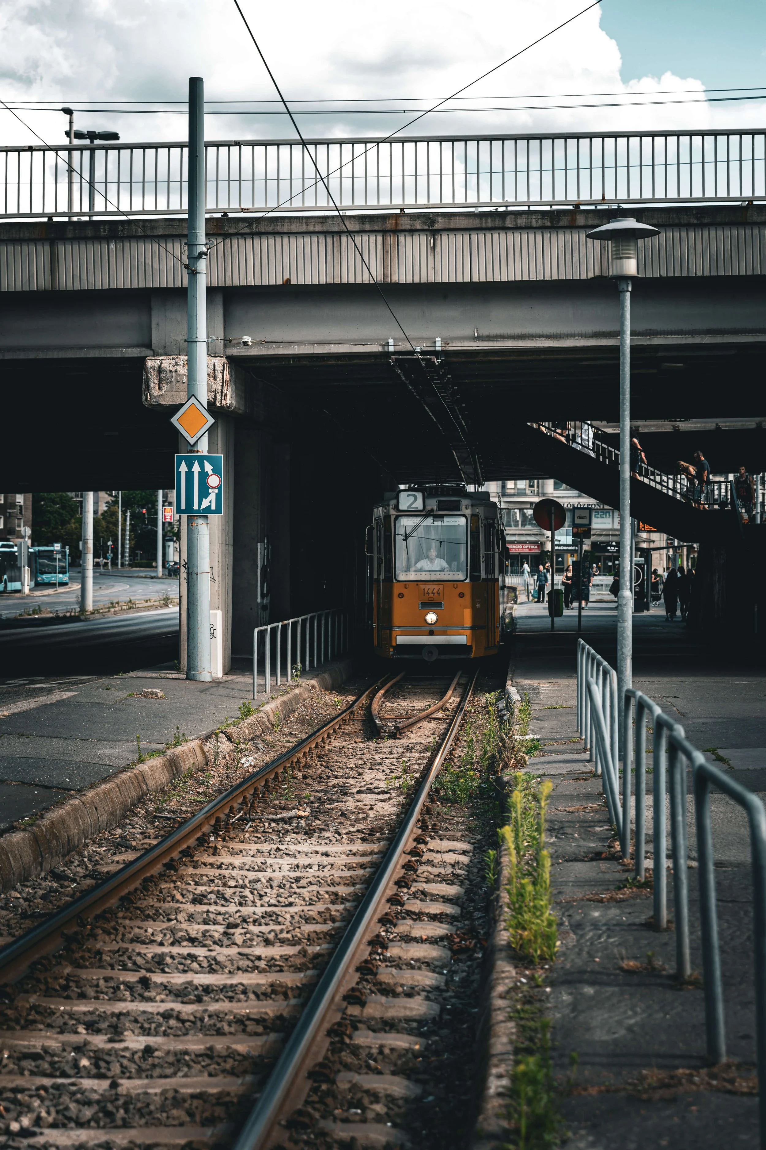 An urban scene with a yellow tram moving on tracks under a bridge, pedestrians using a staircase, and street signs including one with three upward arrows and another with a yellow diamond shape, under a partly cloudy sky.
