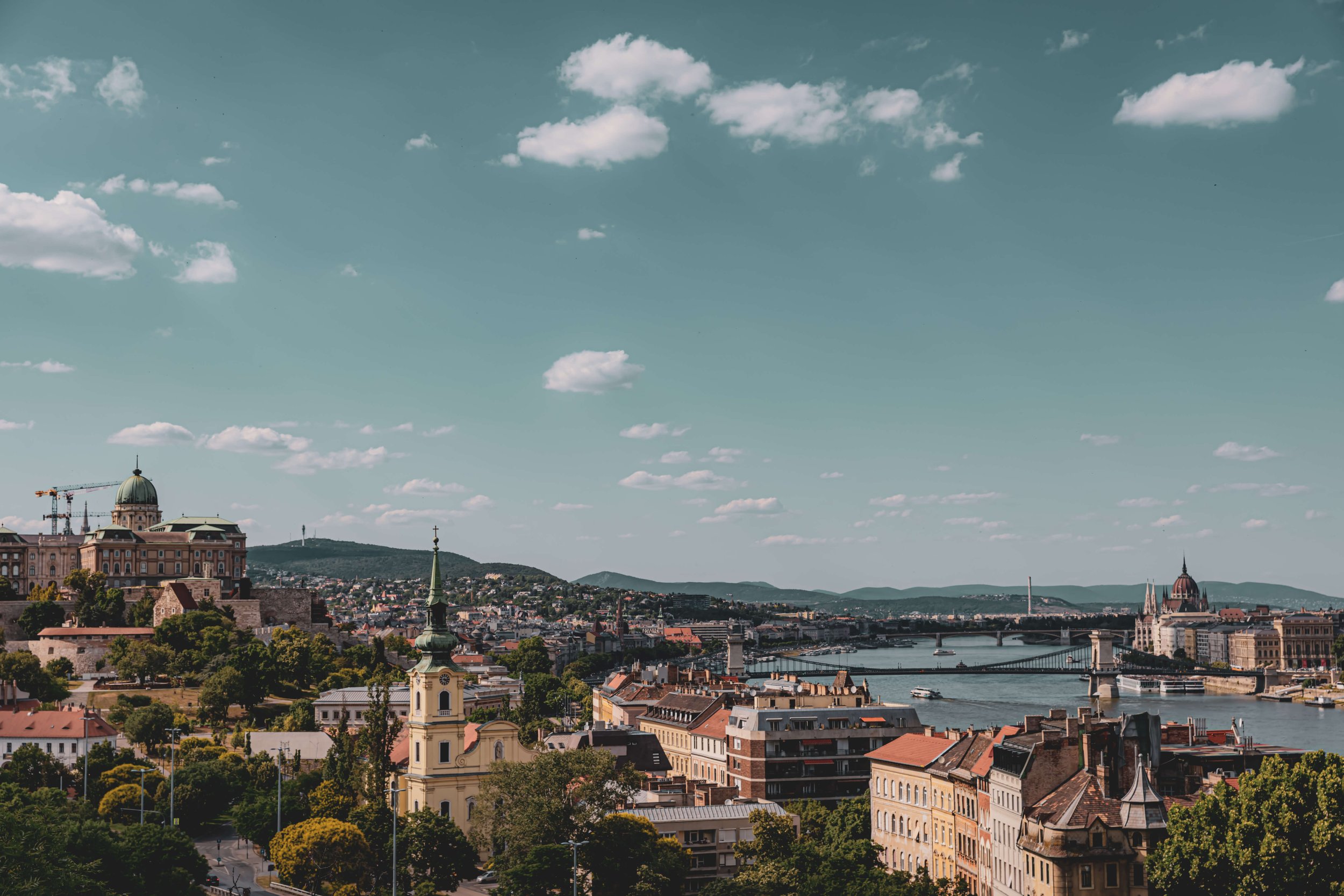 Panoramic view of a city skyline with historic buildings, churches, a bridge across a river, and distant hills under a partly cloudy sky.