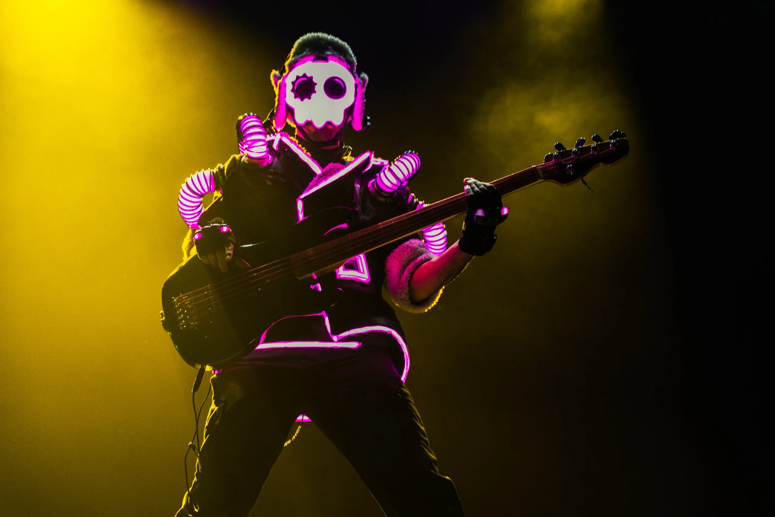 A performer wearing a skull-shaped mask with pink and white lighting, playing an electric bass guitar on stage with yellow stage lighting and a dark background.