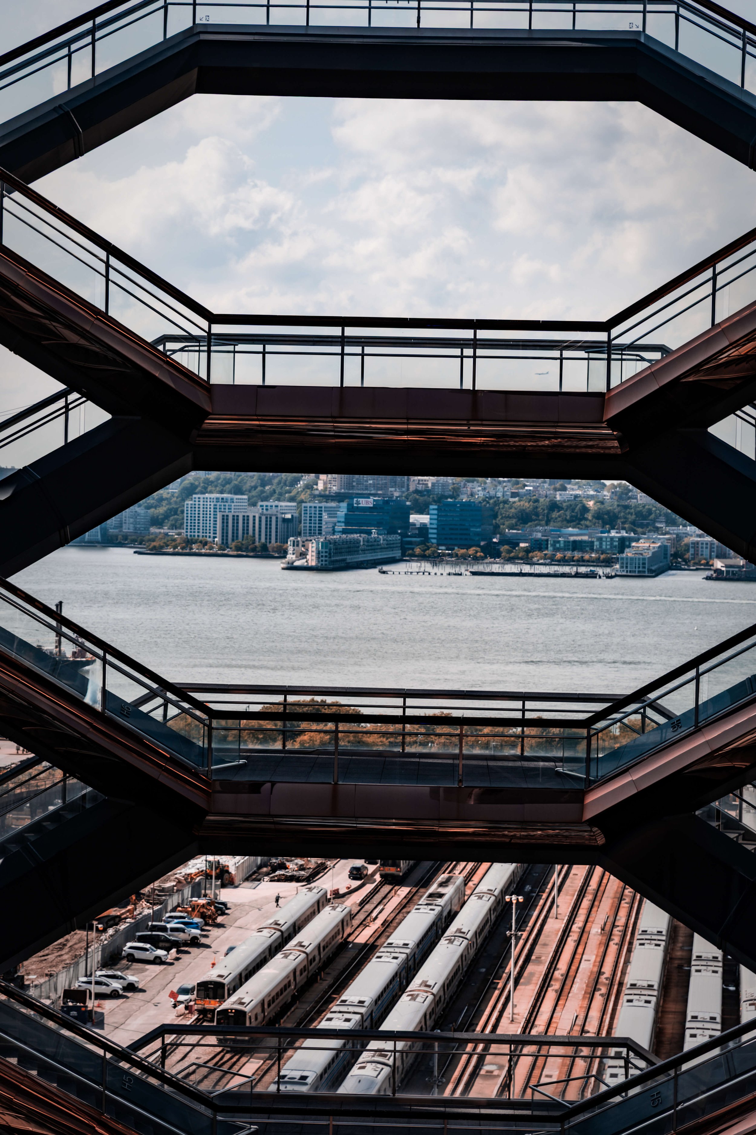 View of a cityscape through a geometric multi-level glass and metal walkway. The city has modern buildings across a body of water, with a train yard and parked trains below.