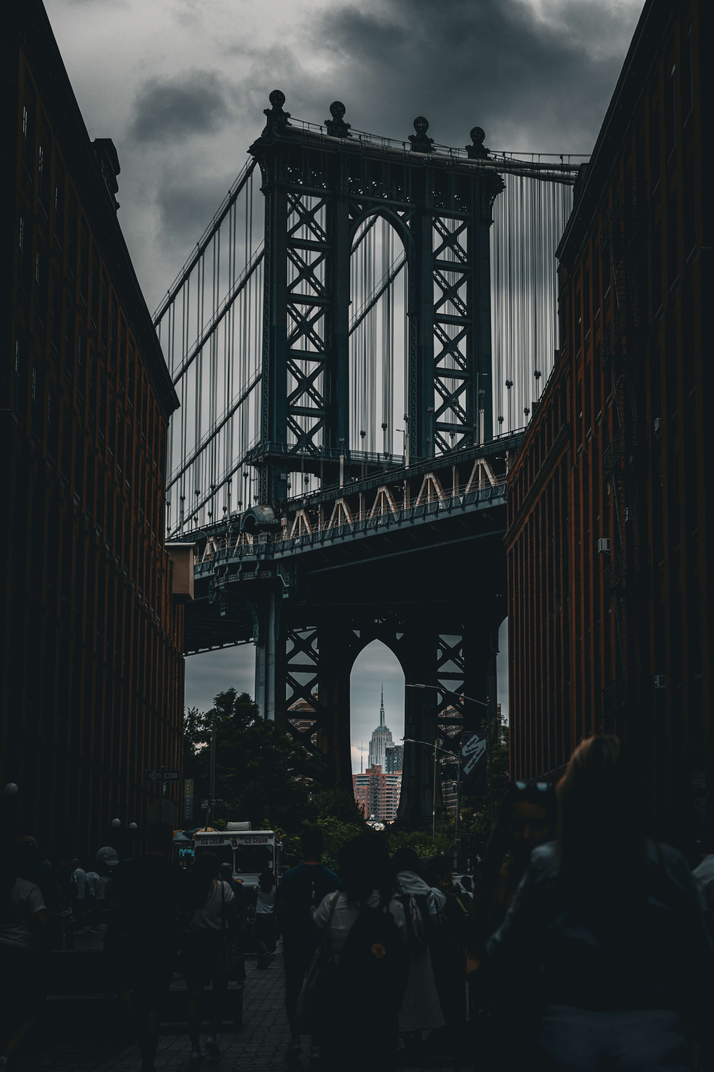 A view of the Manhattan Bridge in New York City with the Empire State Building in the background, taken from a street with people walking and buildings on either side.