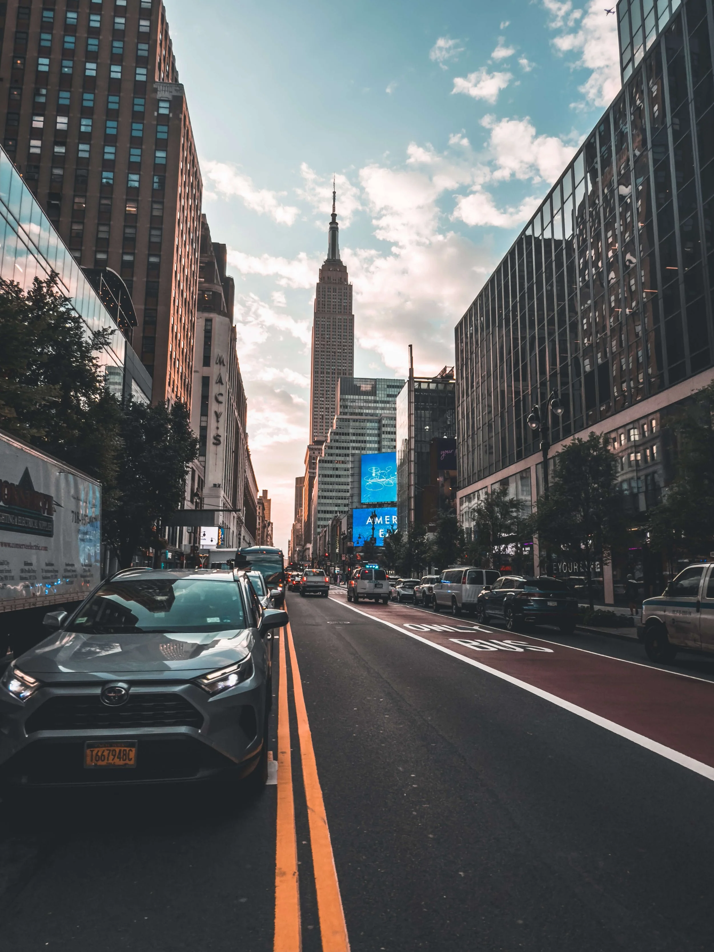 City street in New York City with cars parked along the curb, and the Empire State Building in the background during sunset.