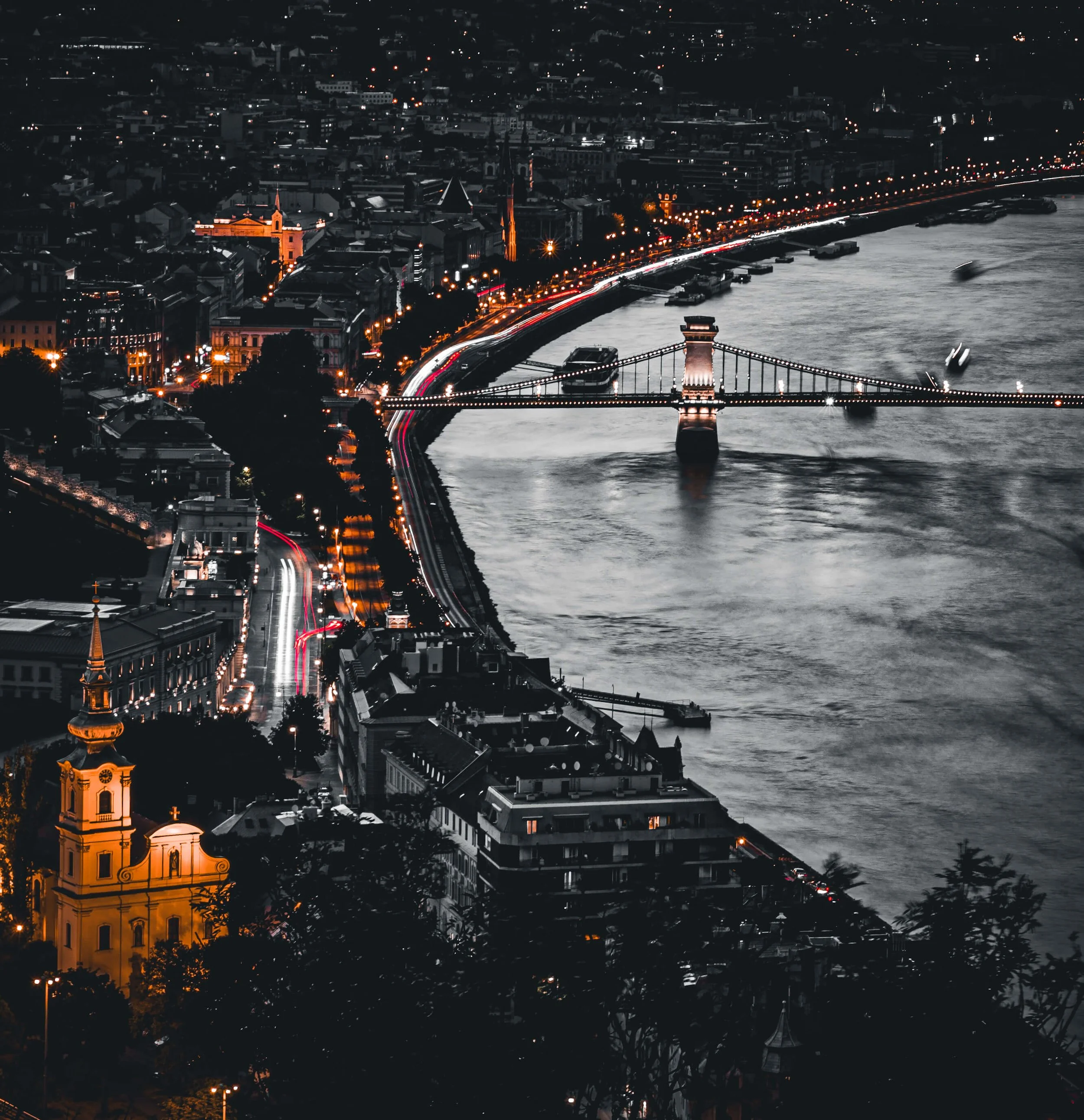 Nighttime aerial view of a city with illuminated streets, historic buildings, and a river with boats. A bridge crosses the river, with city lights and a partly visible church with a bright orange tower in the foreground.