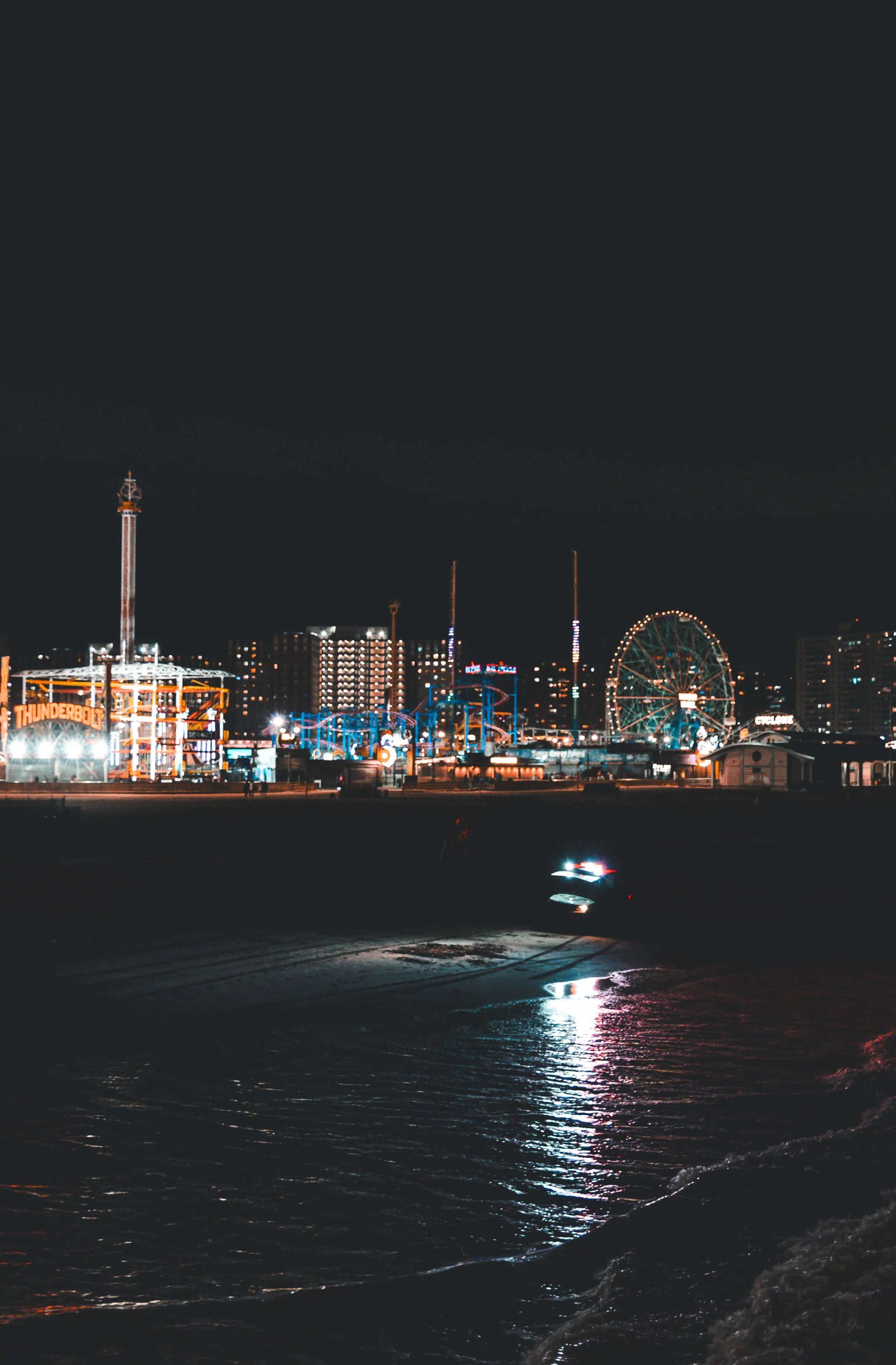 Nighttime scene of an amusement park with a Ferris wheel, roller coasters, and rides illuminated by colorful lights, reflected on the water in the foreground.