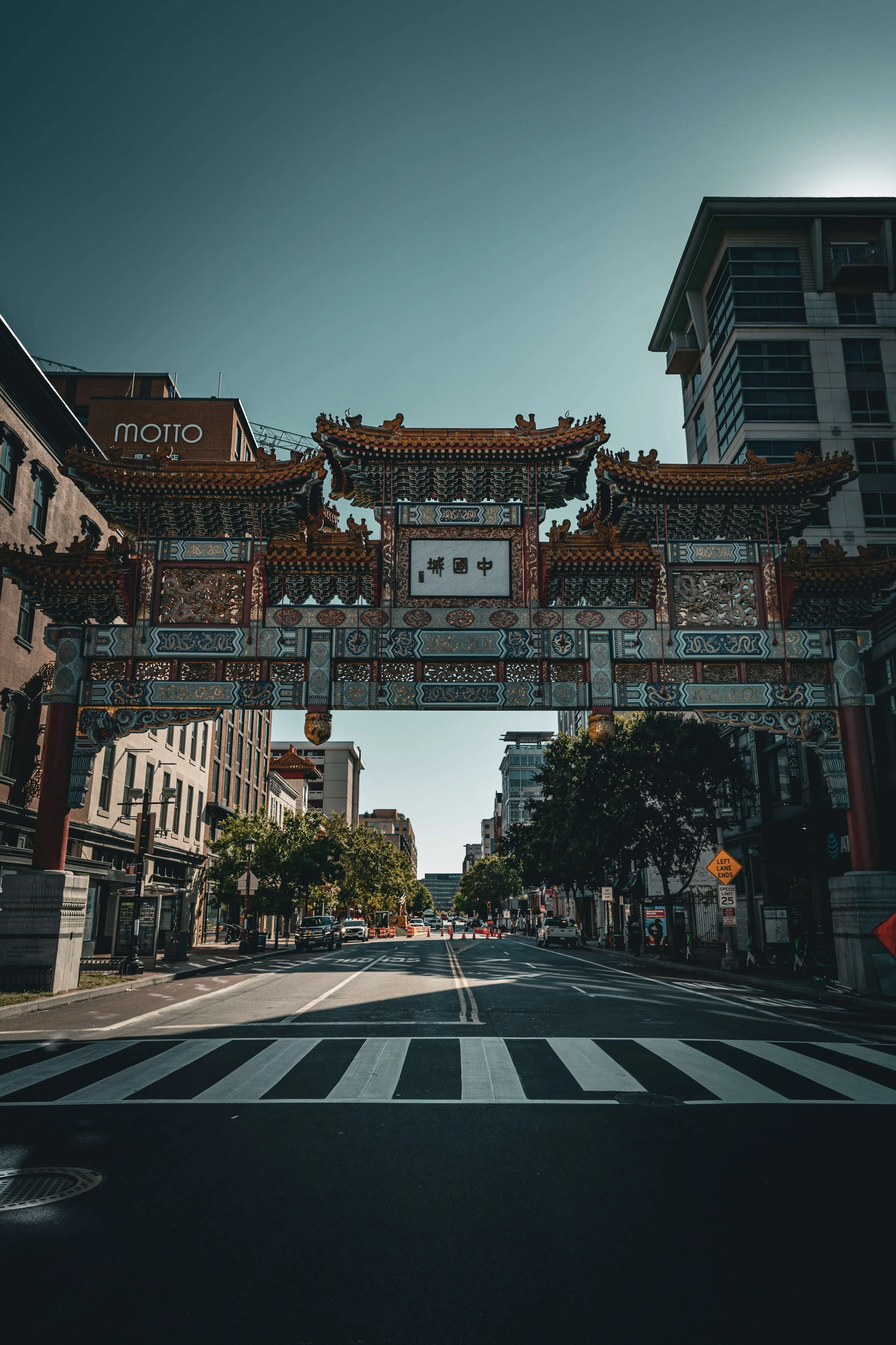 Traditional Chinese gateway archway over a city street with buildings and trees, under a clear sky.