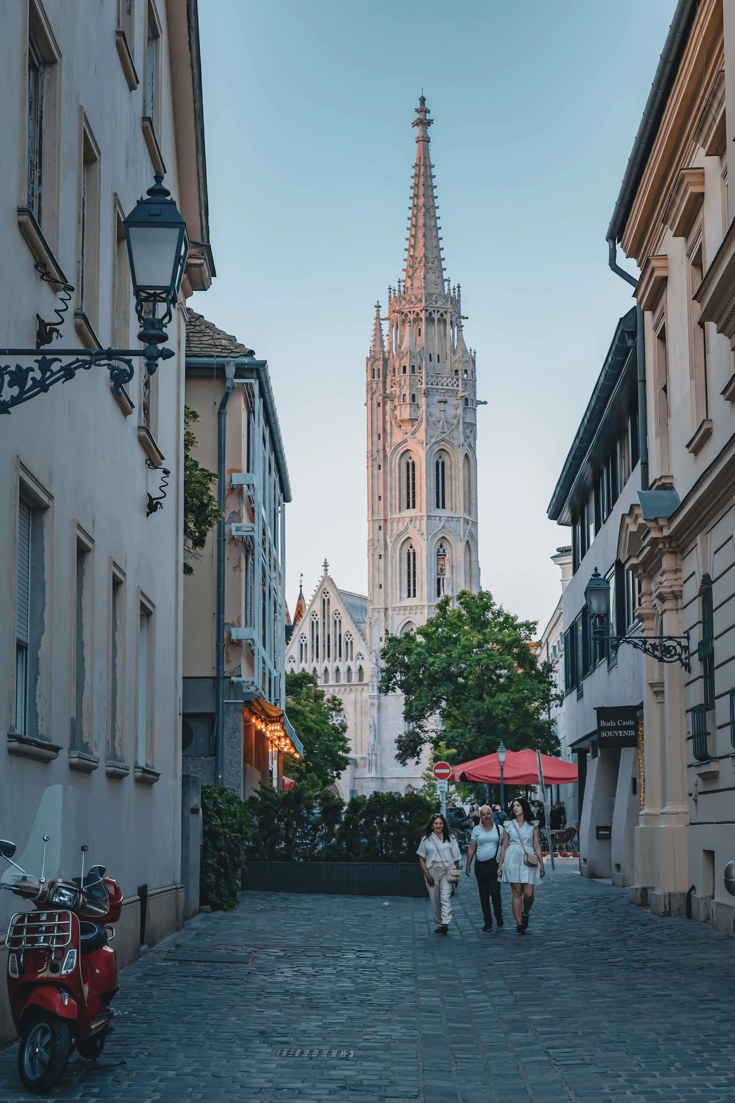 People walking down a cobblestone street lined with historic buildings, with a tall church steeple in the background.