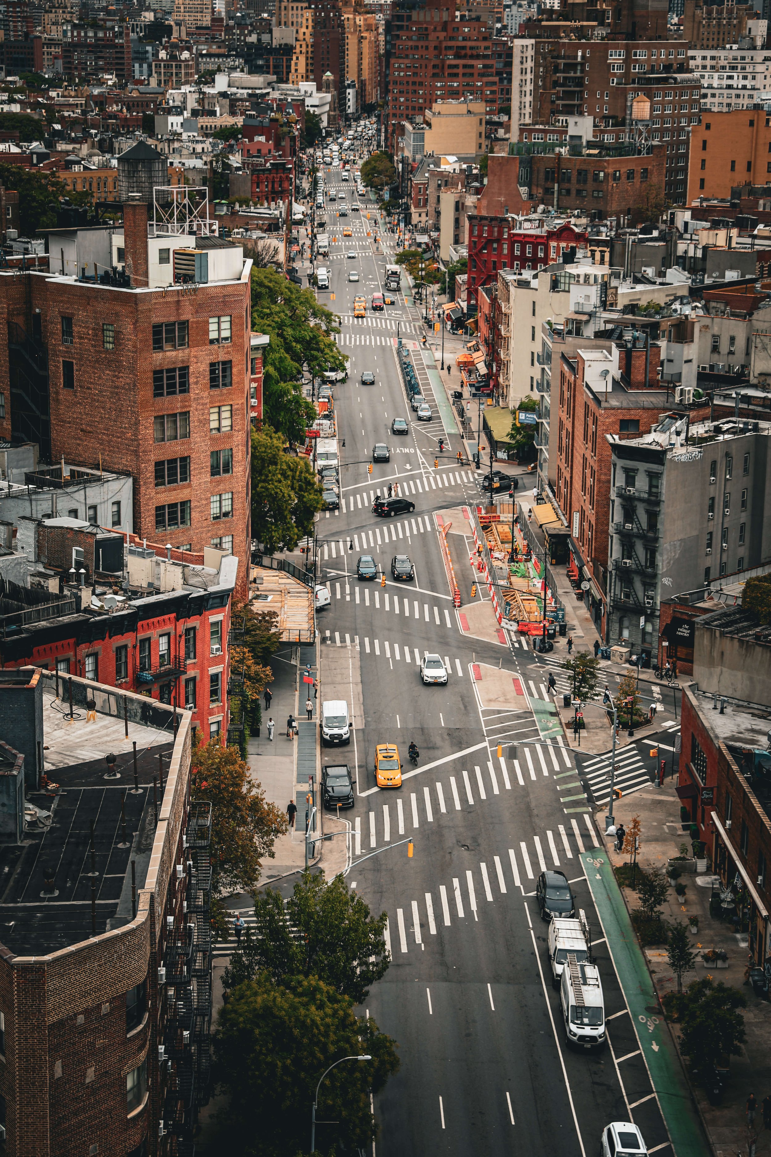 Aerial view of a city street with cars, buses, and pedestrians, surrounded by mid-rise buildings and trees.