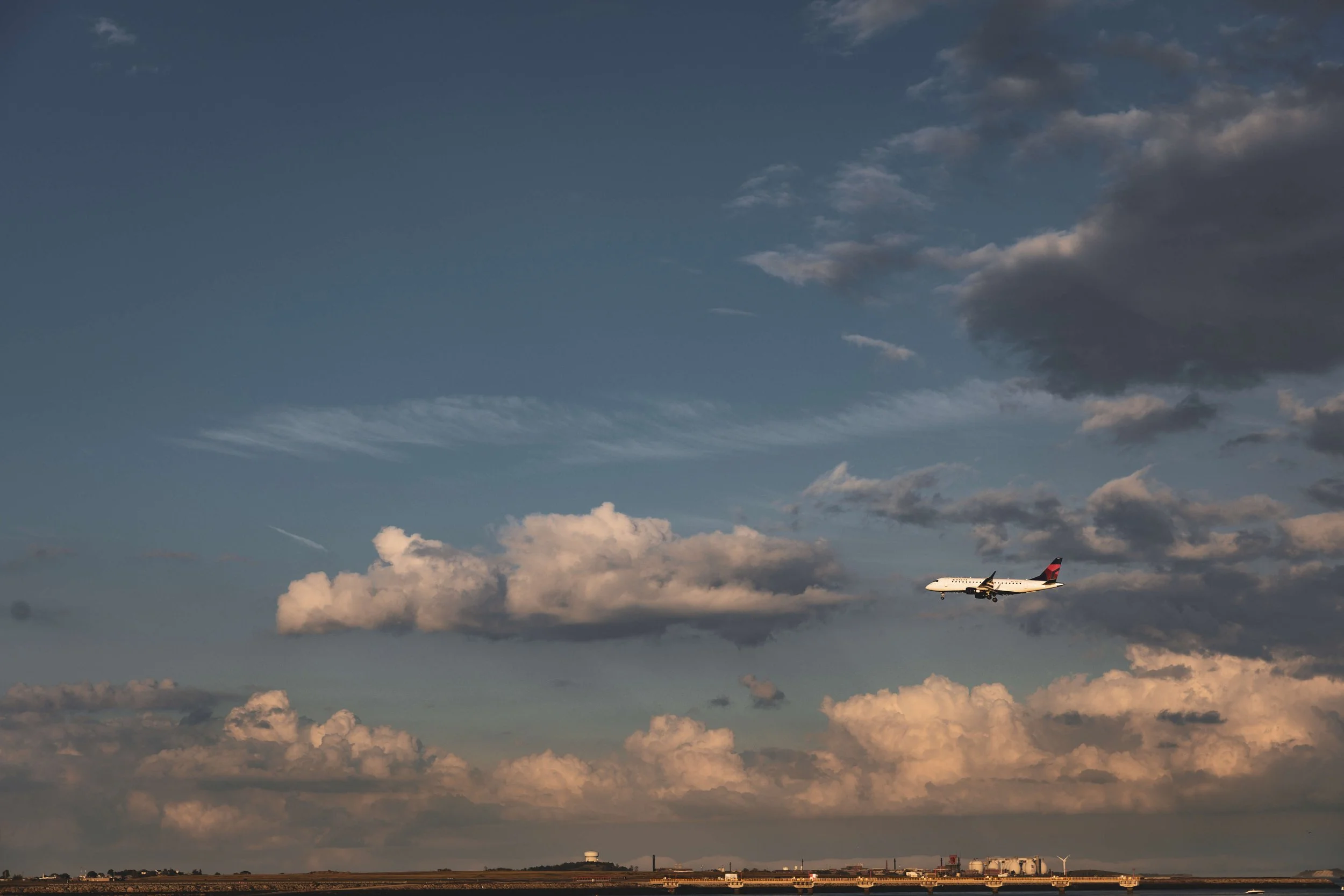 An airplane flying through a partly cloudy sky over a landscape with industrial buildings and wind turbines.