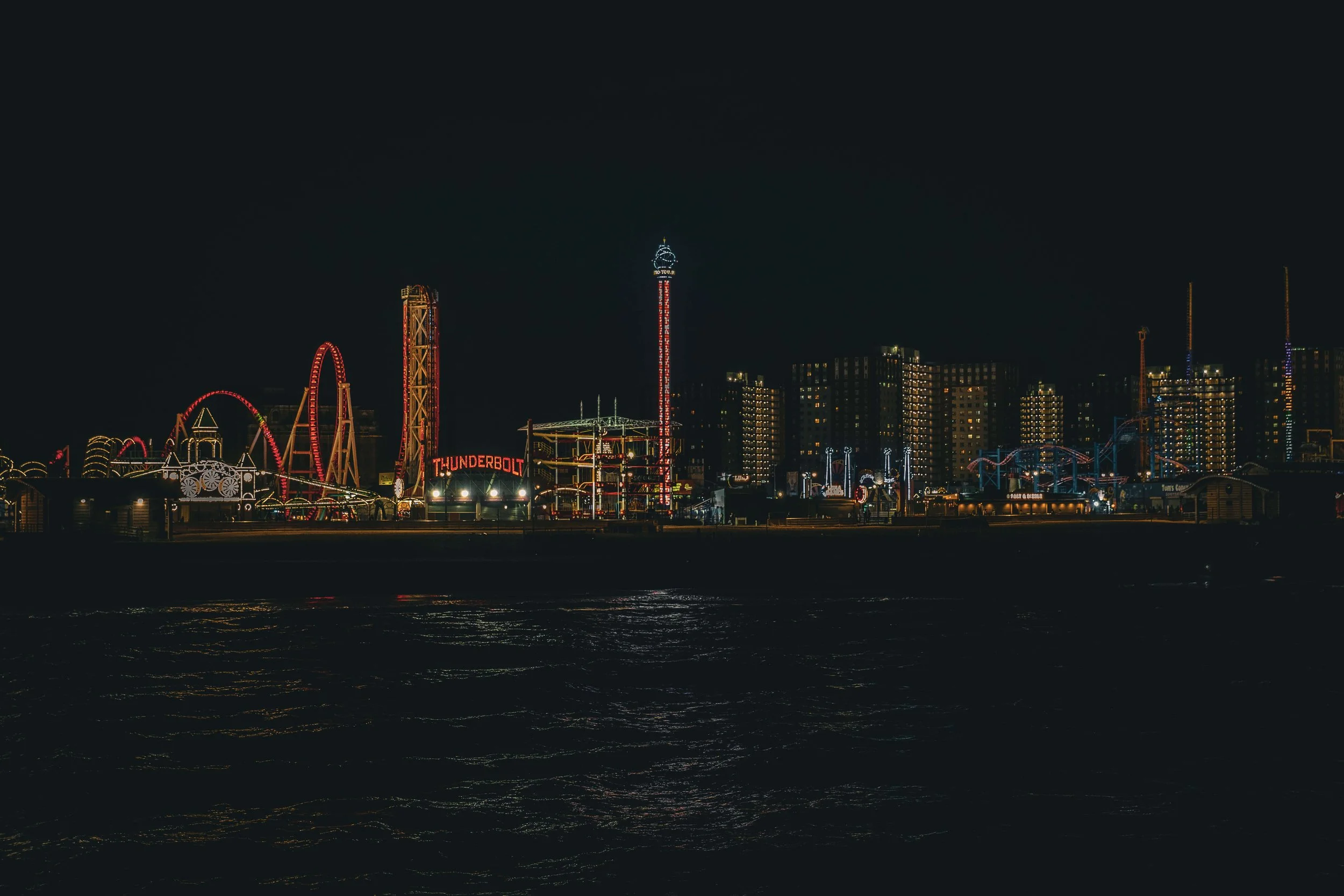 Night view of an amusement park with illuminated rides and attractions, including a roller coaster, drop tower, and the sign 'Thunderbolt,' along a waterfront with dark water in the foreground and high-rise buildings in the background.