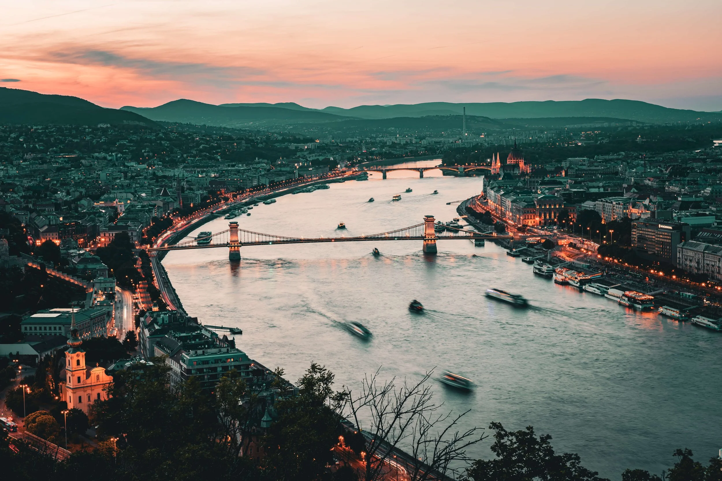 A cityscape at dusk featuring a river with boats, bridges, illuminated buildings including a cathedral, and a skyline with lush green hills in the background.