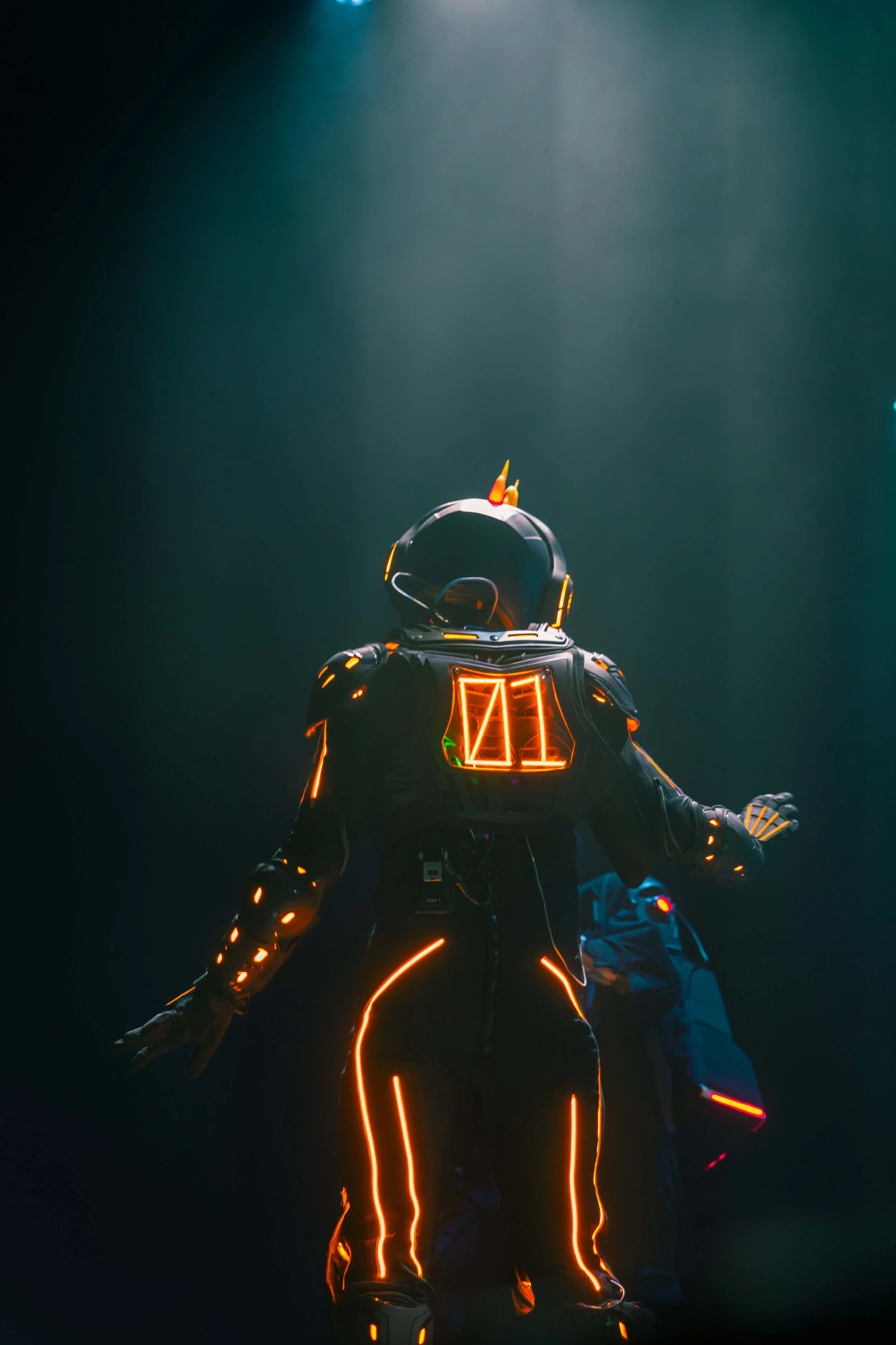 Person in a futuristic, neon-lit suit standing in a dark environment with glowing orange accents and a robot or another person in the background.