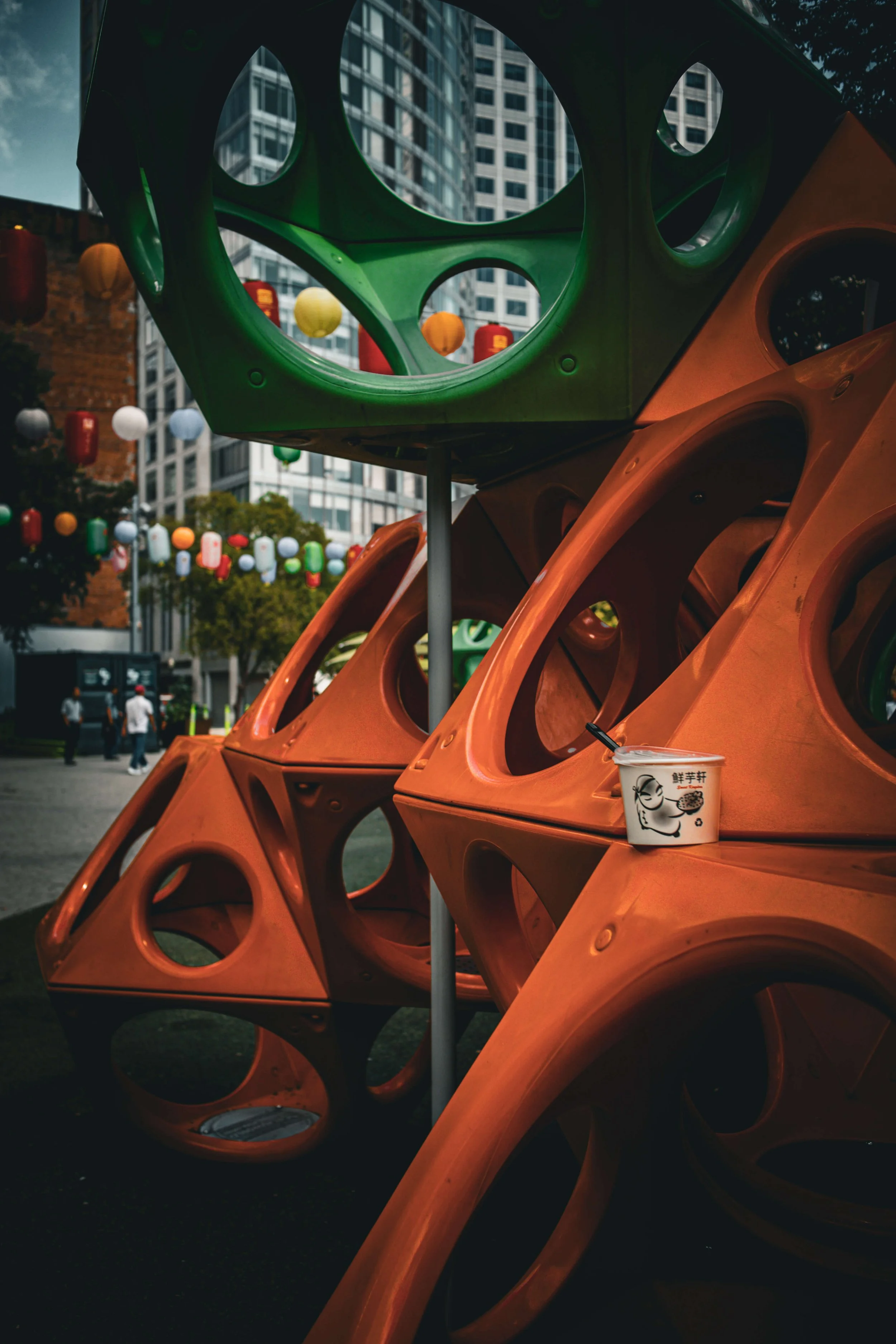 Colorful geometric outdoor art installation with green and orange structures, paper lanterns hanging in the background, and a small cup with an illustration placed on one of the structures, in an urban setting.