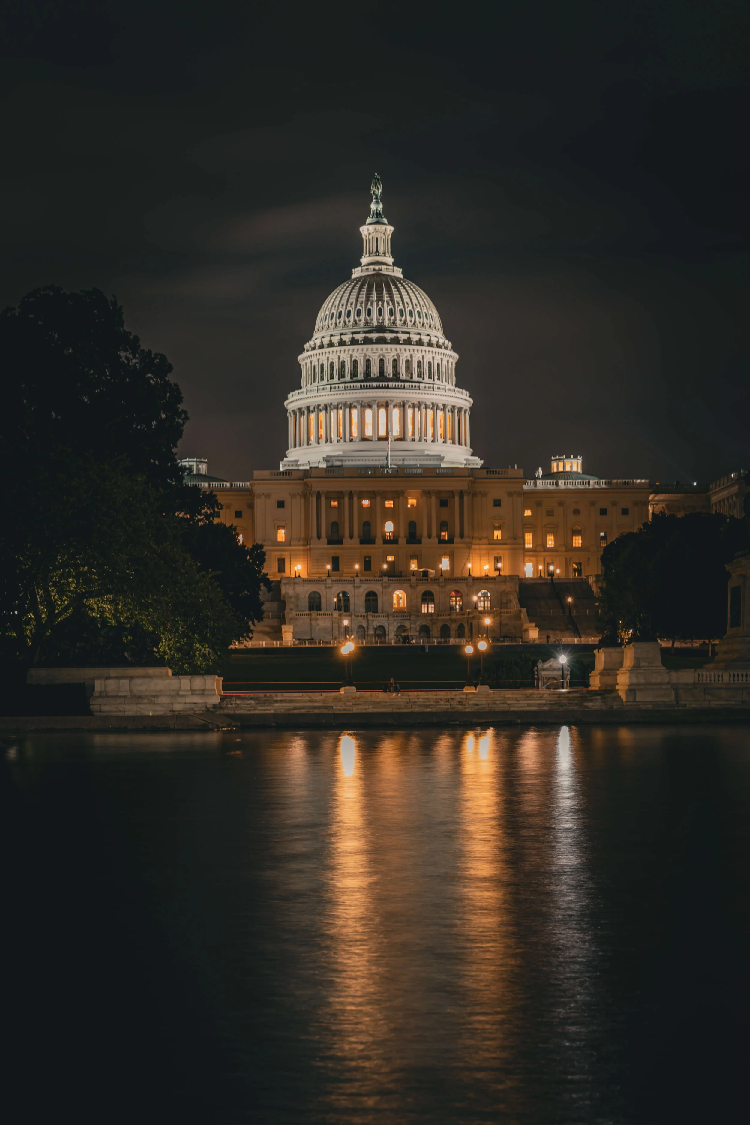 Night view of the United States Capitol building illuminated, with reflections on the water in front.