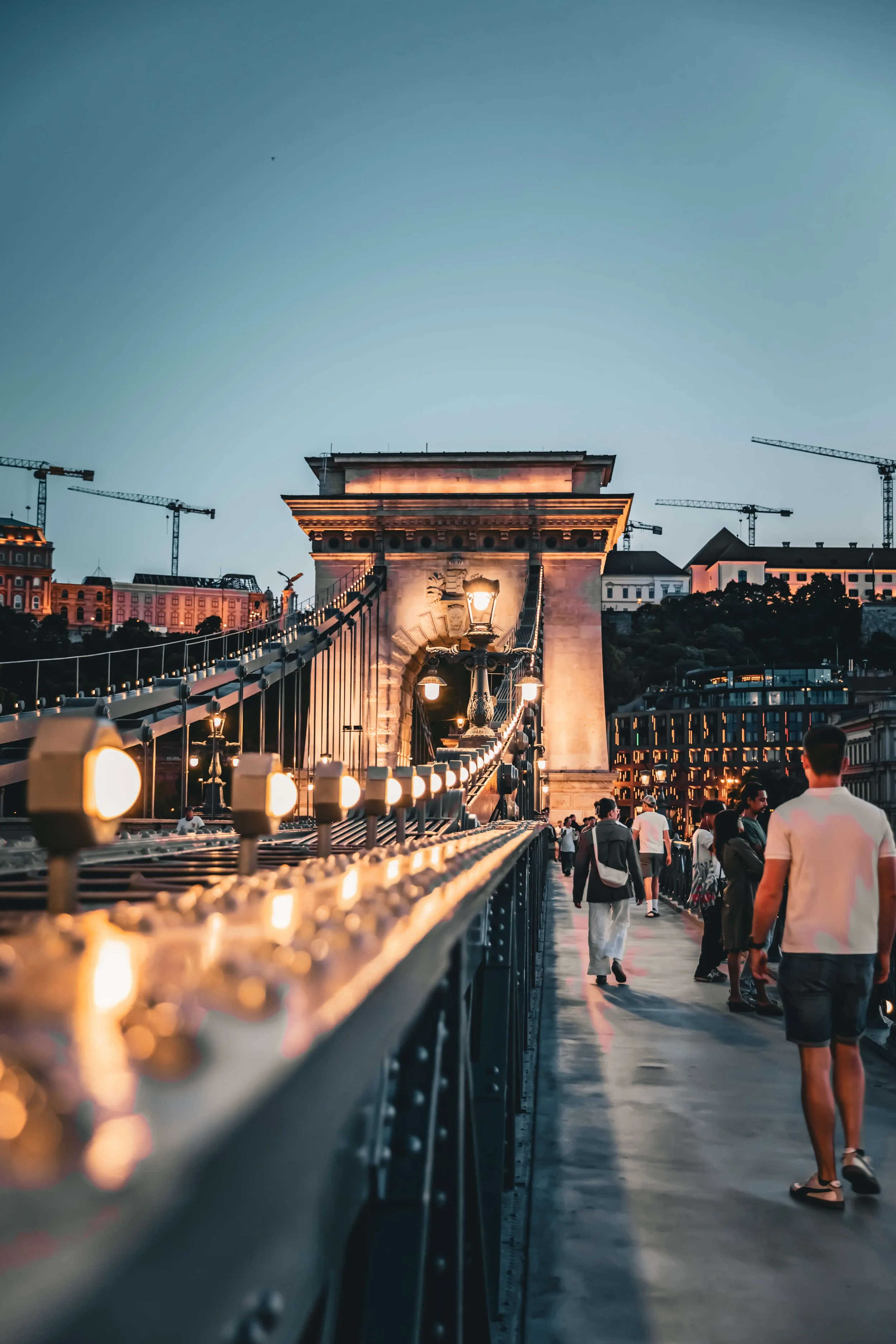 A suspension bridge illuminated with lights during twilight, with people walking on the pedestrian walkway and construction cranes in the background.