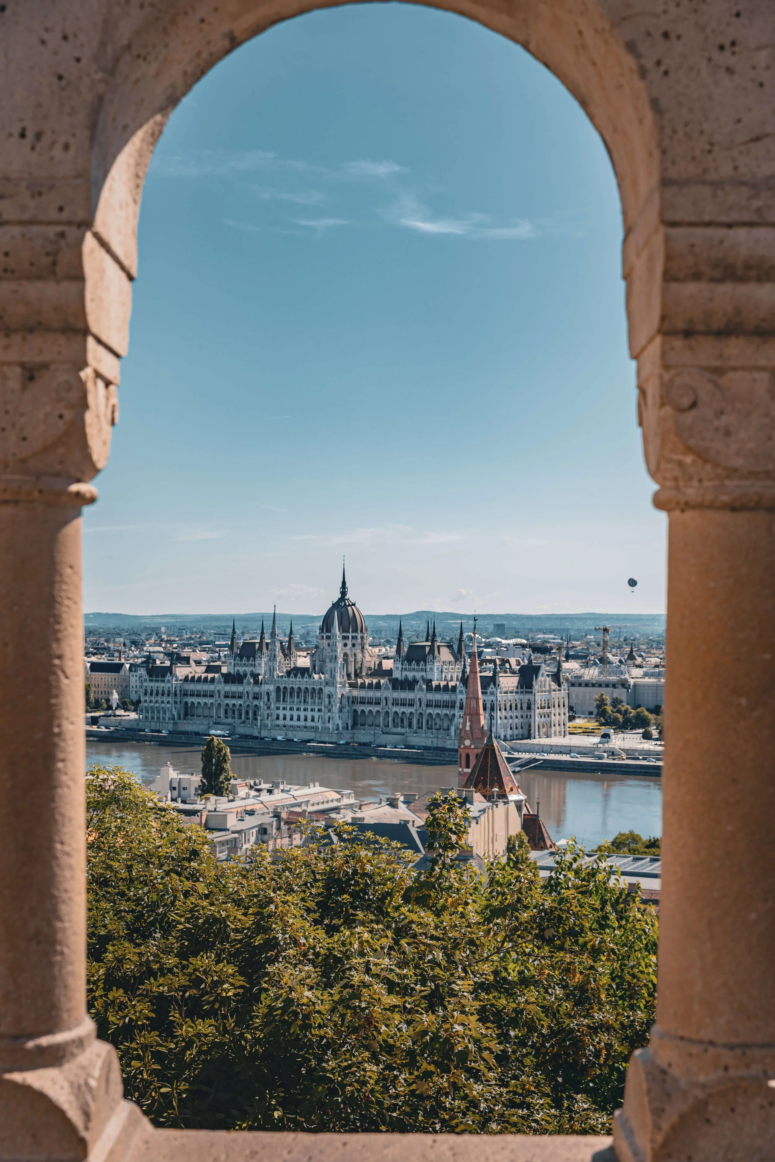 View of Budapest, Hungary, seen through an arched stone window, featuring the Parliament building along the Danube River, with a clear blue sky overhead.