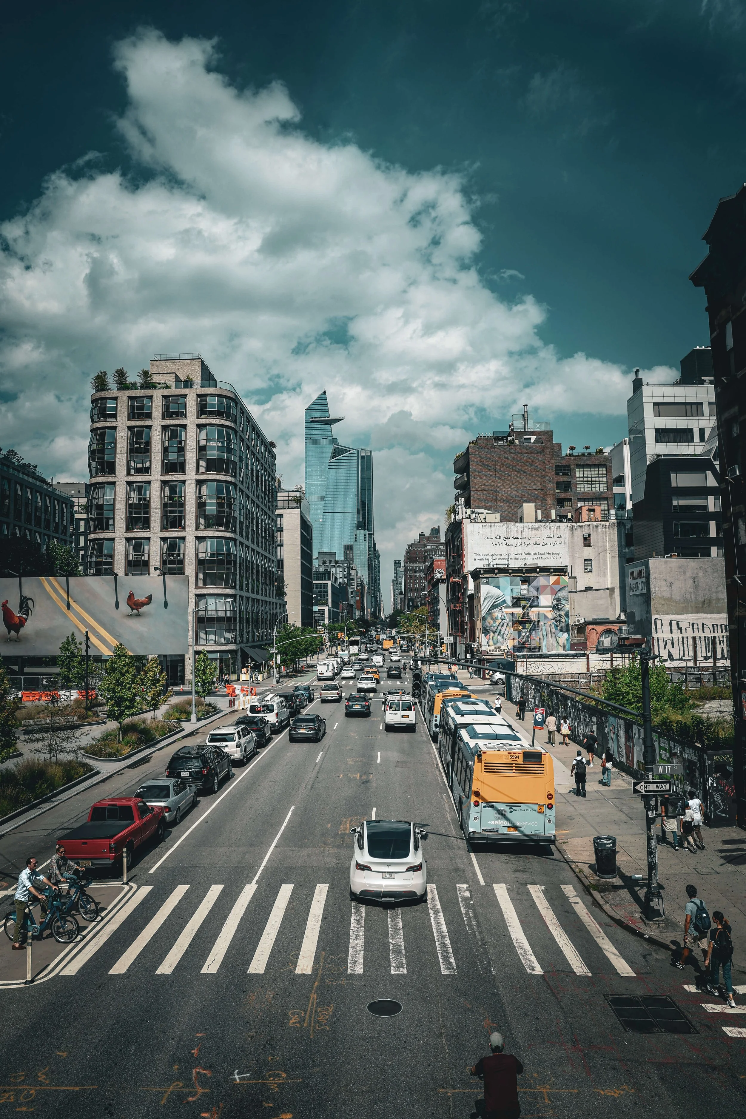 Urban city street with cars, buses, and pedestrians, tall buildings, and a partly cloudy sky.
