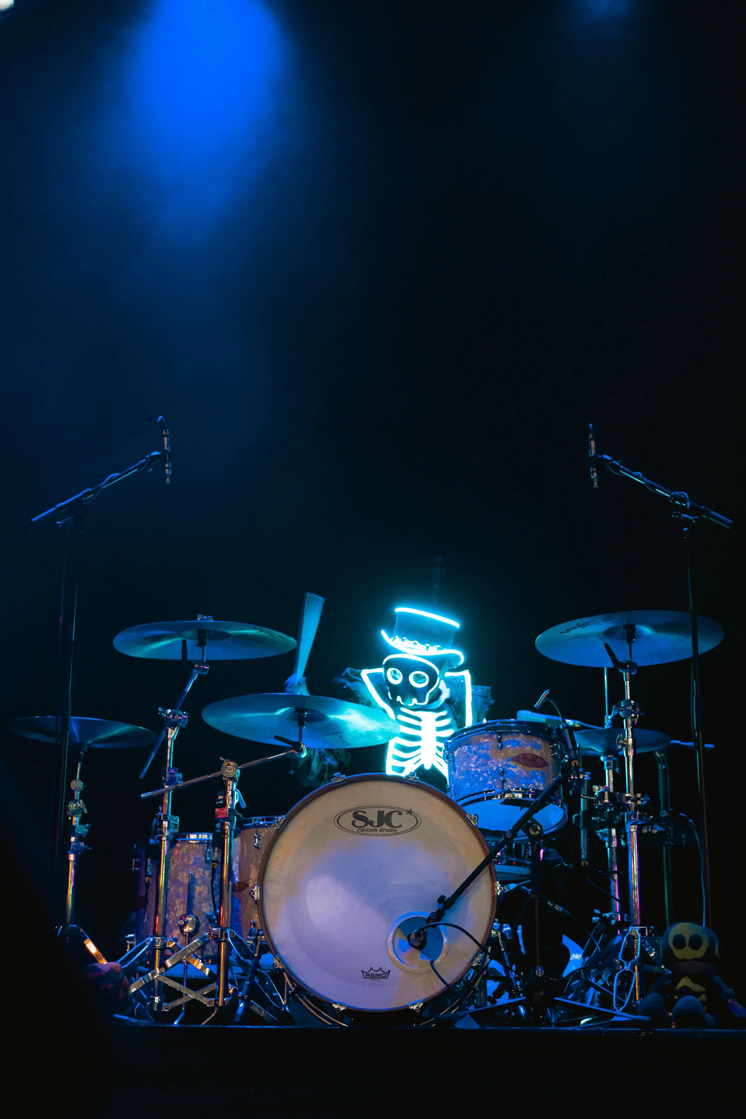 Stage with a drum kit illuminated by blue and white lights, featuring a skeleton with a hat in the background
