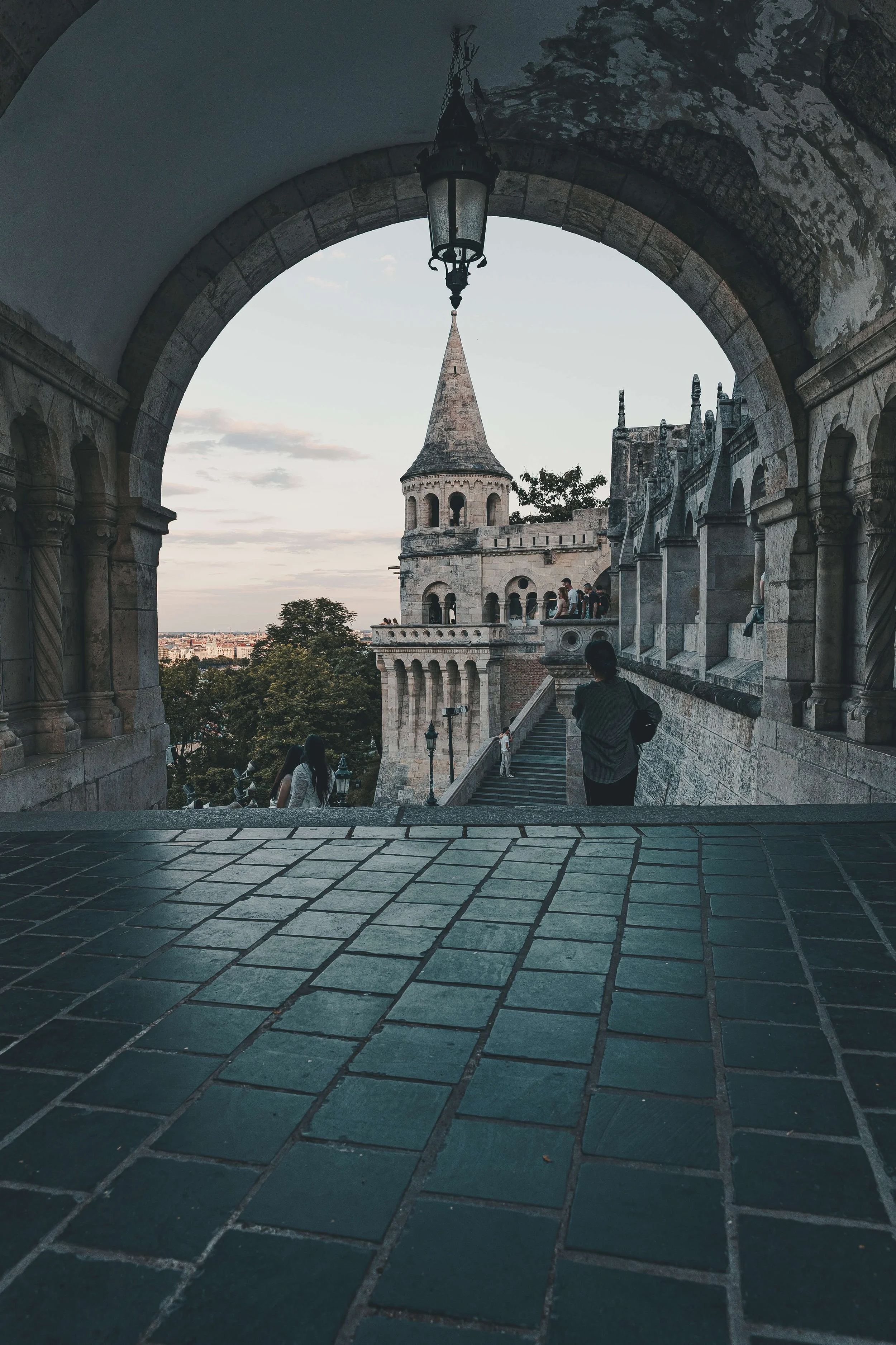 View of a historic castle or palace with a tower featuring a pointed roof, framed by a stone archway. Several people are walking on the stone stairs and balcony area, with cityscape visible in the background during dusk or evening.