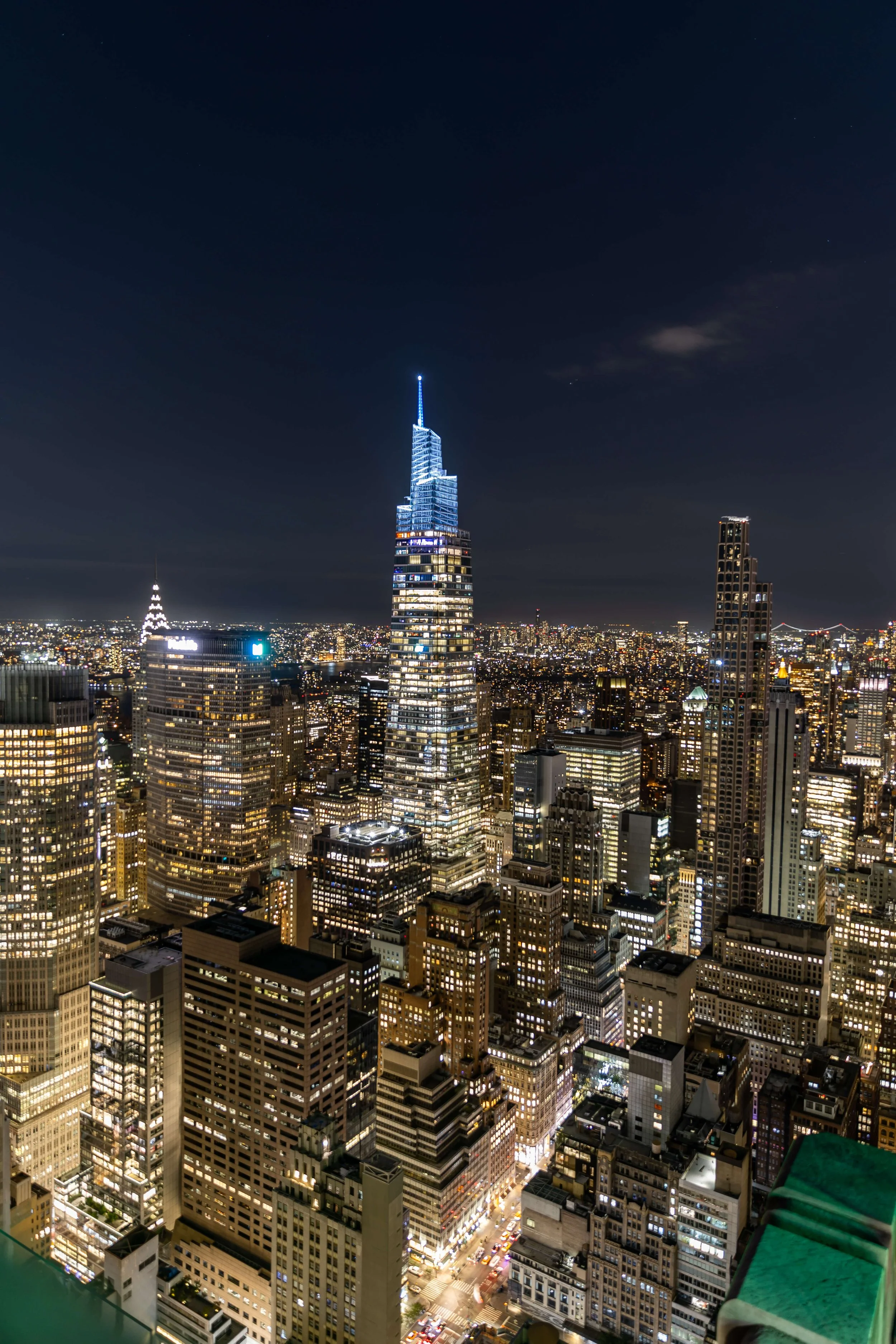 Nighttime cityscape of New York City with illuminated skyscrapers, including a prominent tall building with a spire, viewed from a high vantage point.