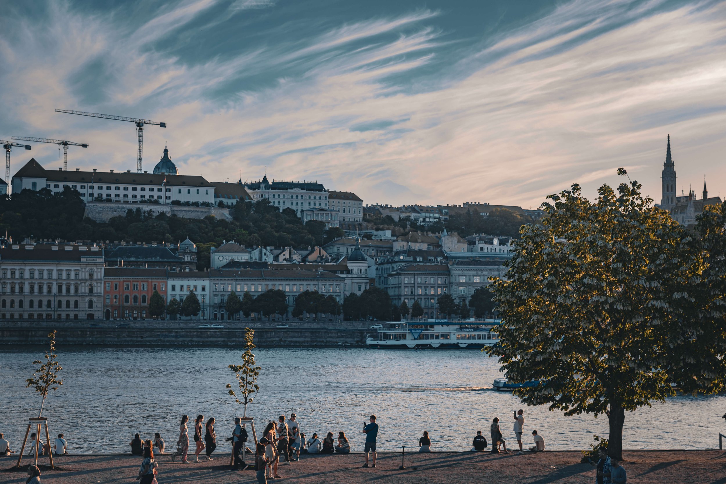 People walking and sitting by a river with a city skyline in the background, including buildings on a hill, construction cranes, and a church spire, with a partly cloudy sky overhead.