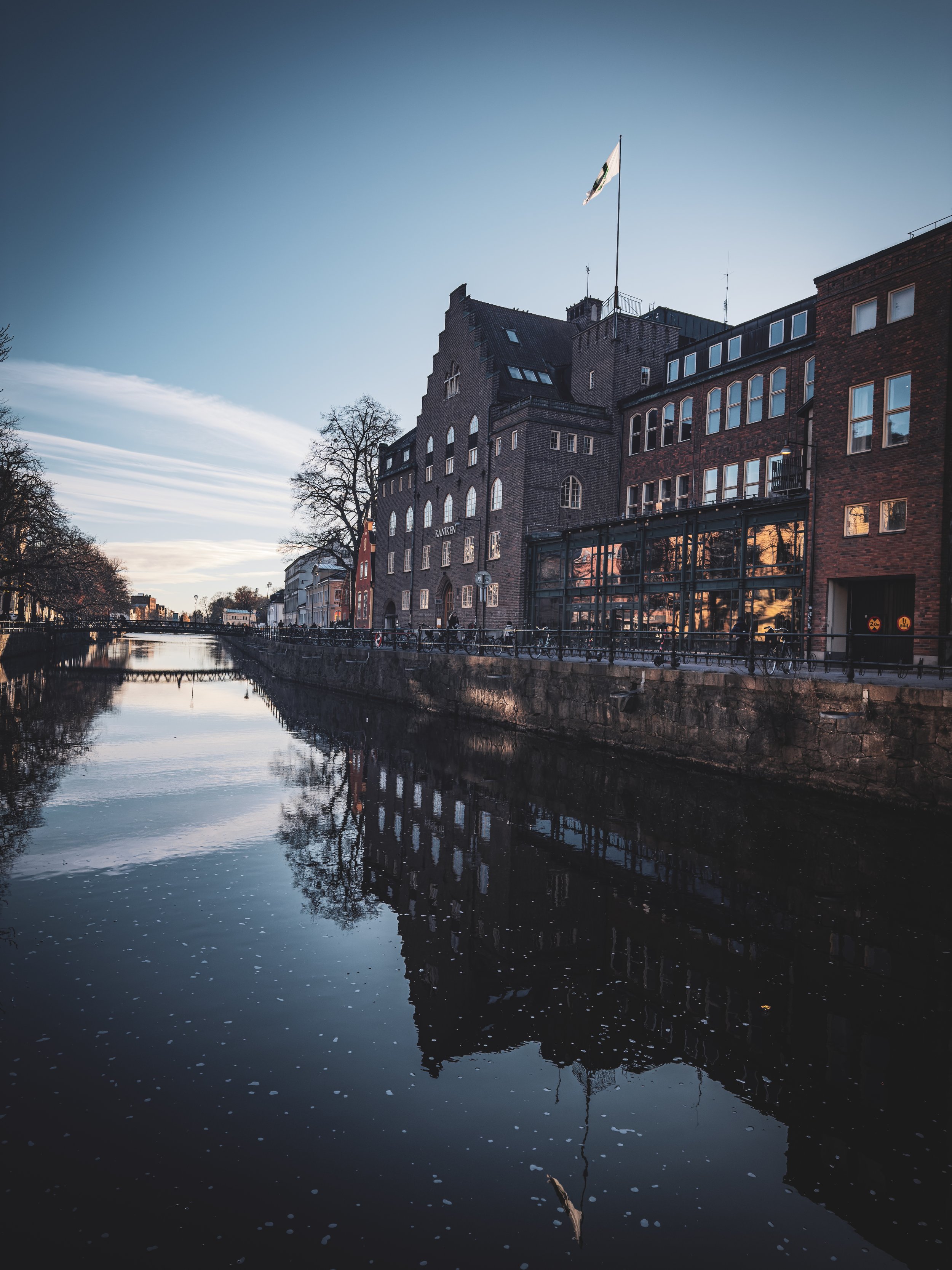 A reflective canal lined with historic brick buildings and bare trees during sunset, under a clear blue sky.