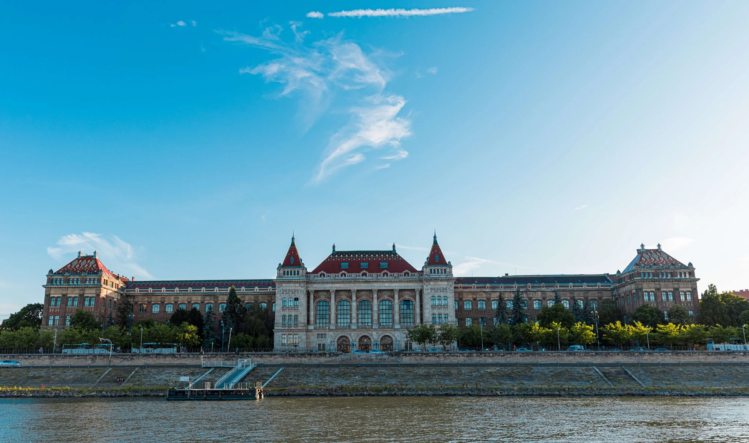 Historic building with a red roof and ornate architecture, situated by a river with green trees in the foreground and a blue sky with clouds above.