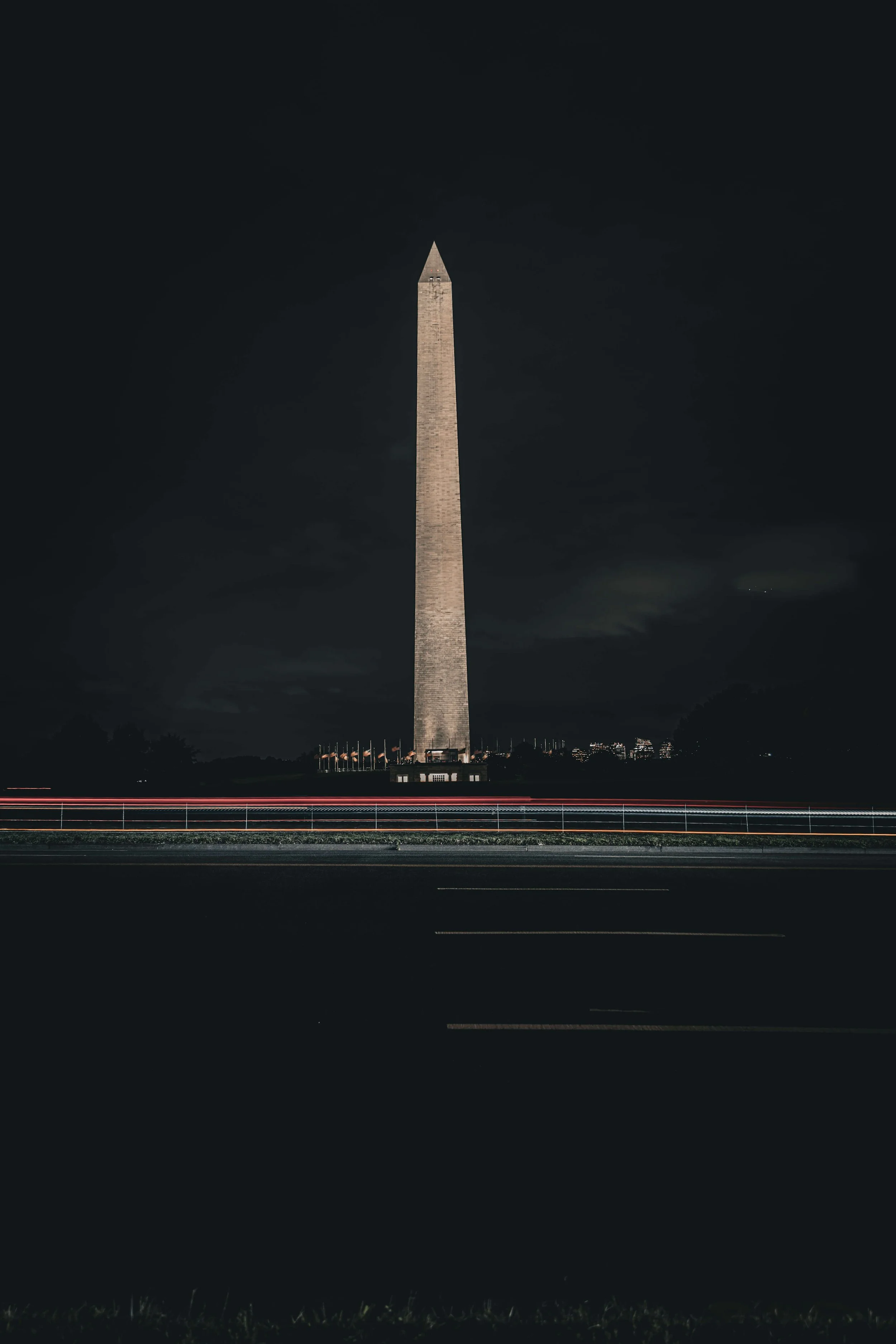 Nighttime view of the Washington Monument in Washington, D.C., with light trails from passing cars in the foreground.
