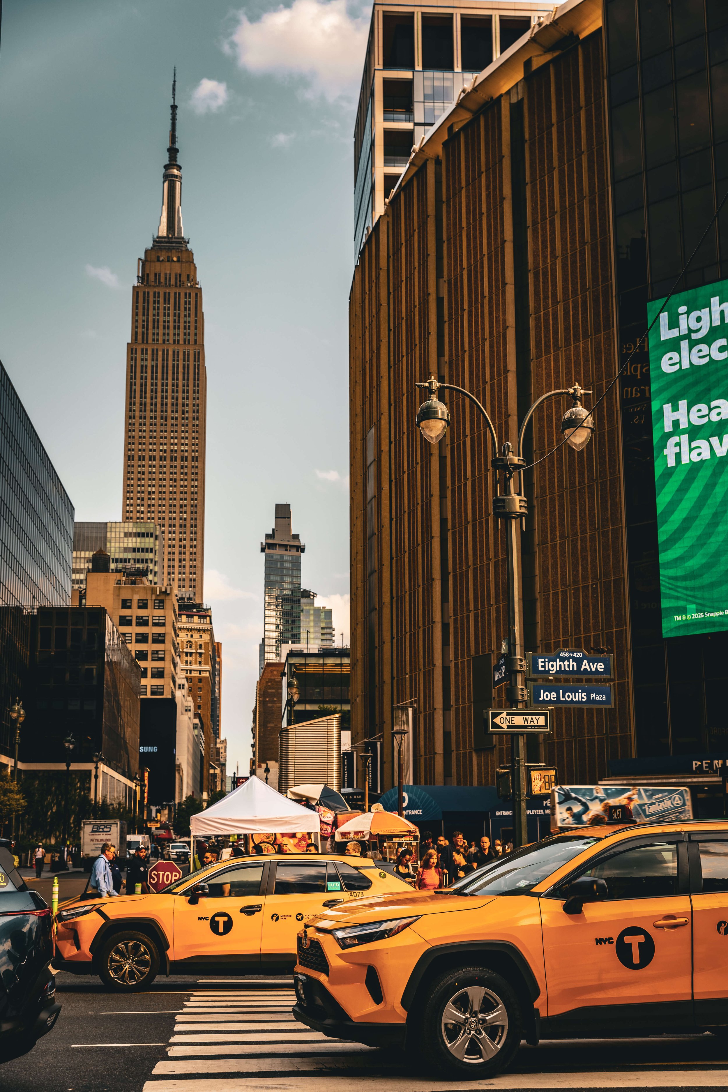 New York City street scene with yellow taxis, pedestrians, tall buildings including the Empire State Building, street signs for Eighth Ave and Joe Louis Plaza, and a digital billboard.