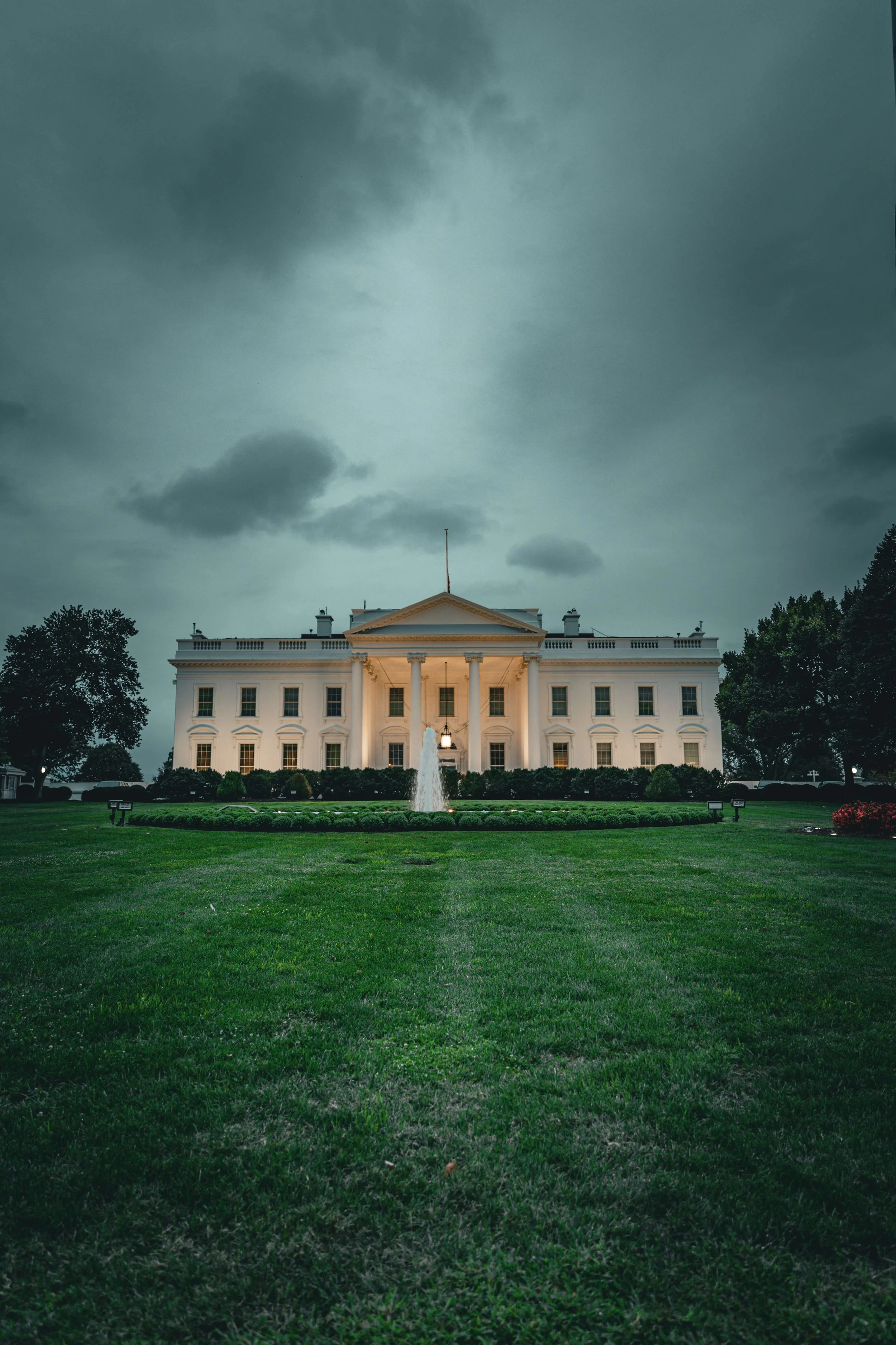 The White House, the official residence and workplace of the President of the United States, under a cloudy sky, with a fountain and green lawn in the foreground.