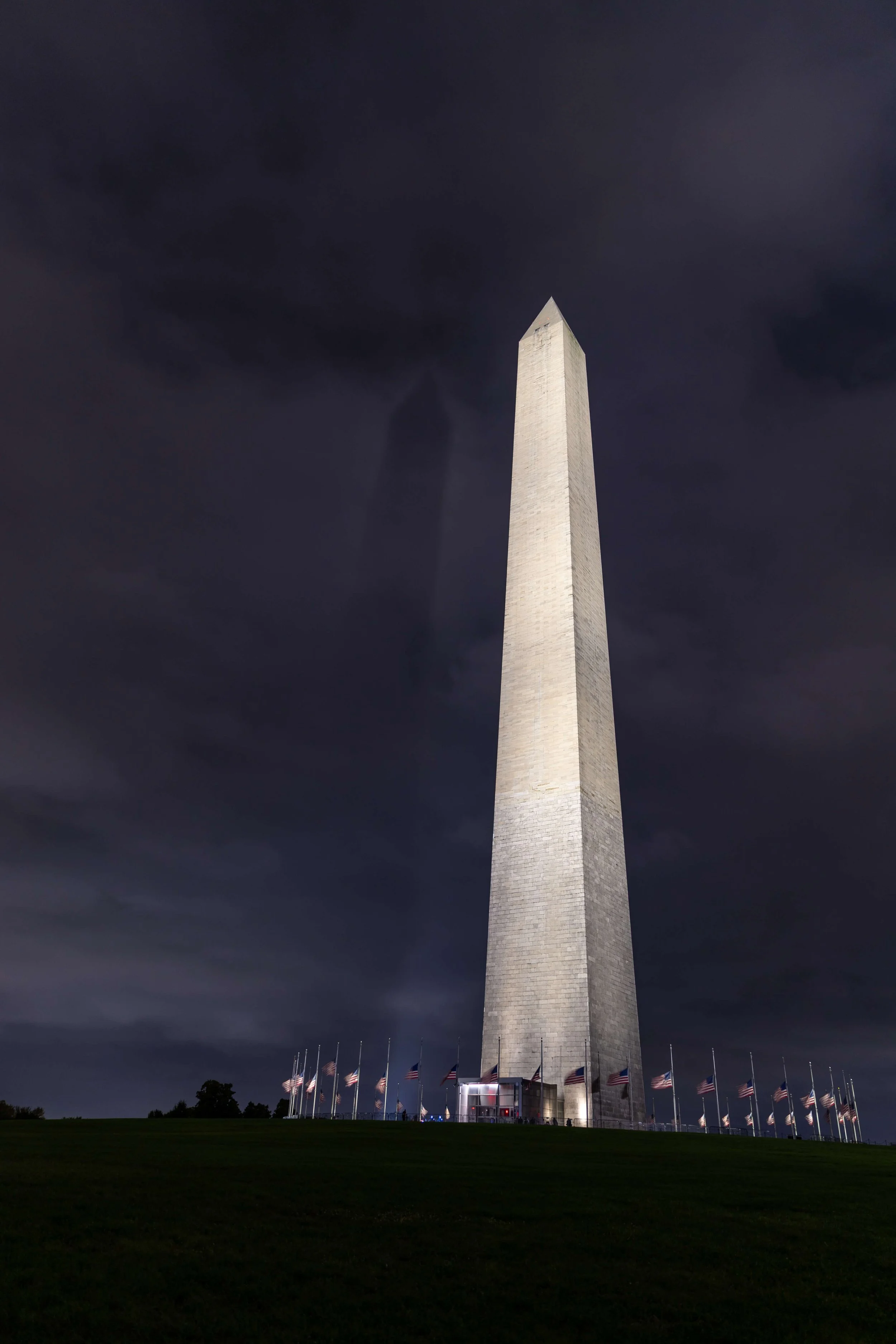 The Washington Monument illuminated at night with American flags surrounding its base, under a dark cloudy sky.