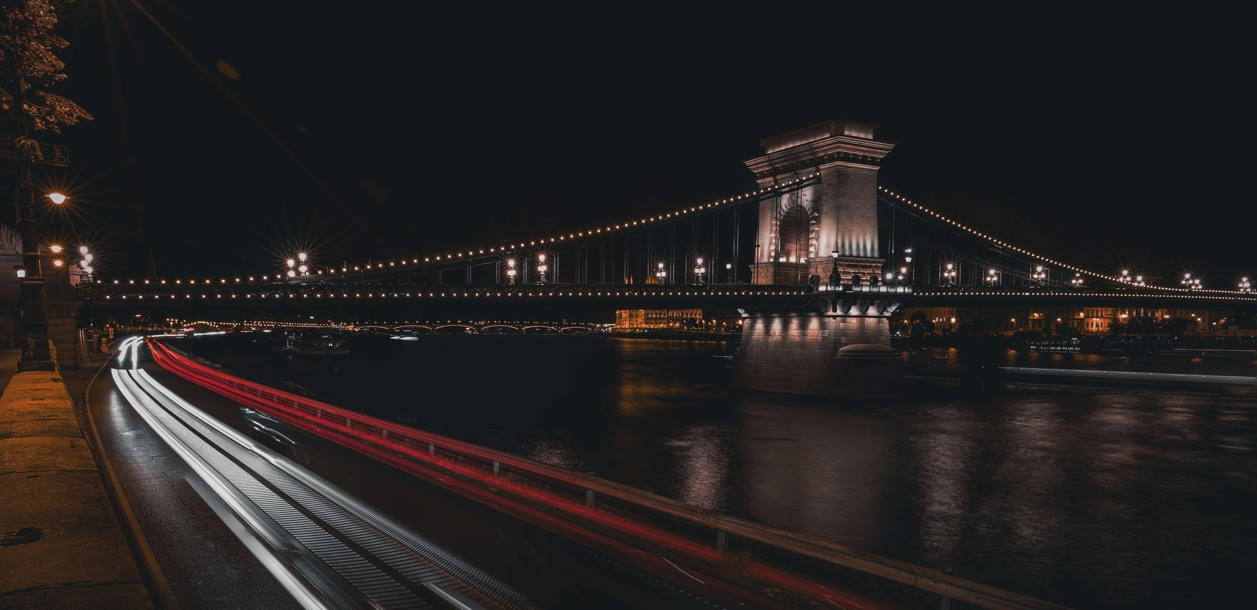 Night view of the illuminated chain bridge over a river in Budapest, with light trails from moving vehicles along the riverside road.