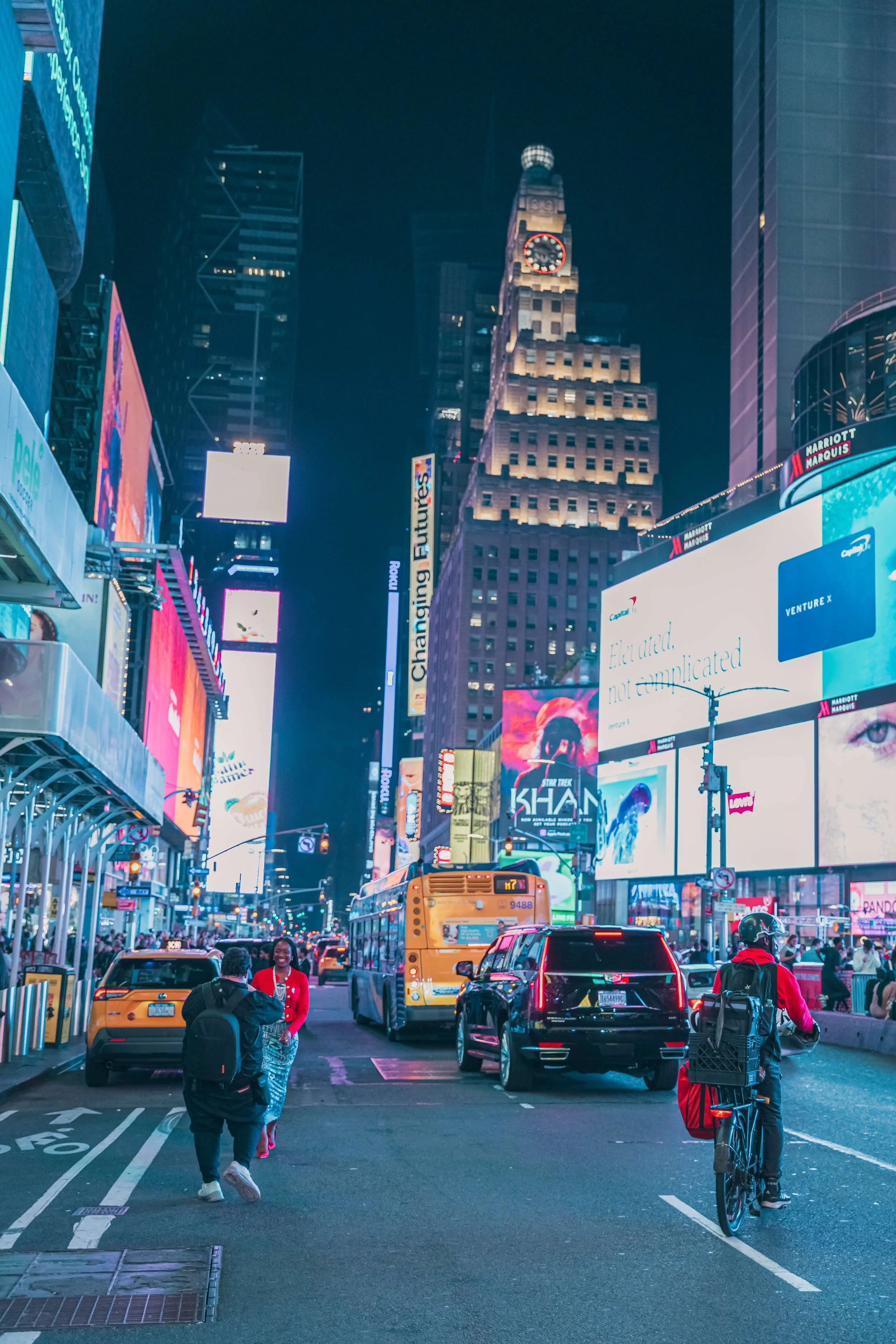 Nighttime scene in Times Square, New York City with bright digital billboards, yellow taxis, a bus, pedestrians, and a cyclist.