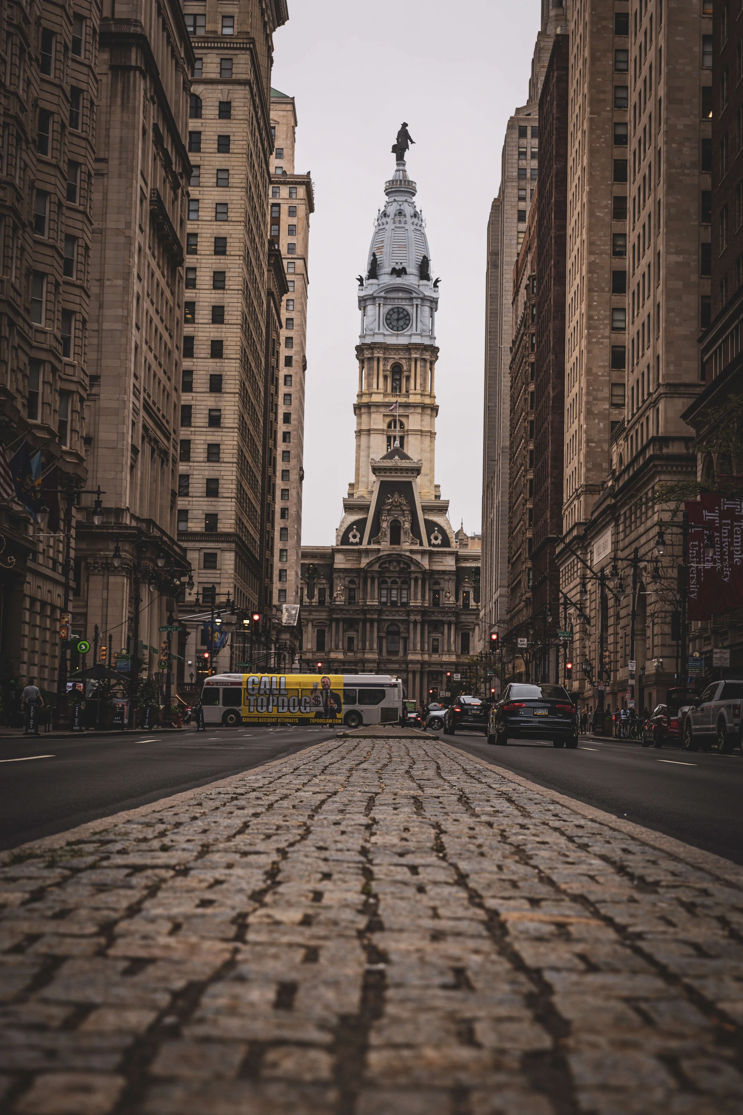 View of Philadelphia City Hall with cars and a bus on the cobblestone street, surrounded by tall buildings and overcast sky.