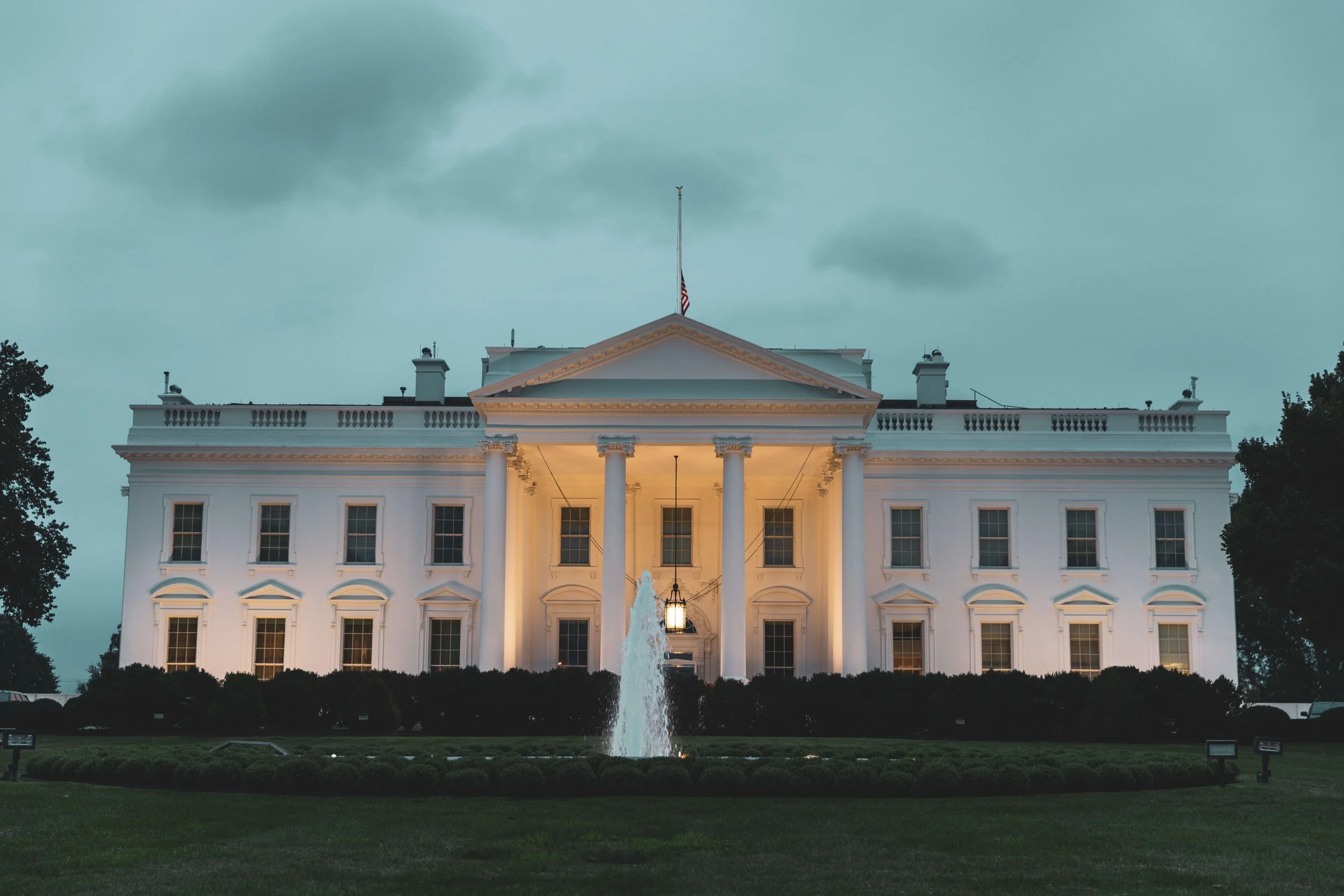 The White House illuminated at dusk with a fountain in front, surrounded by a well-kept lawn and shrubbery.