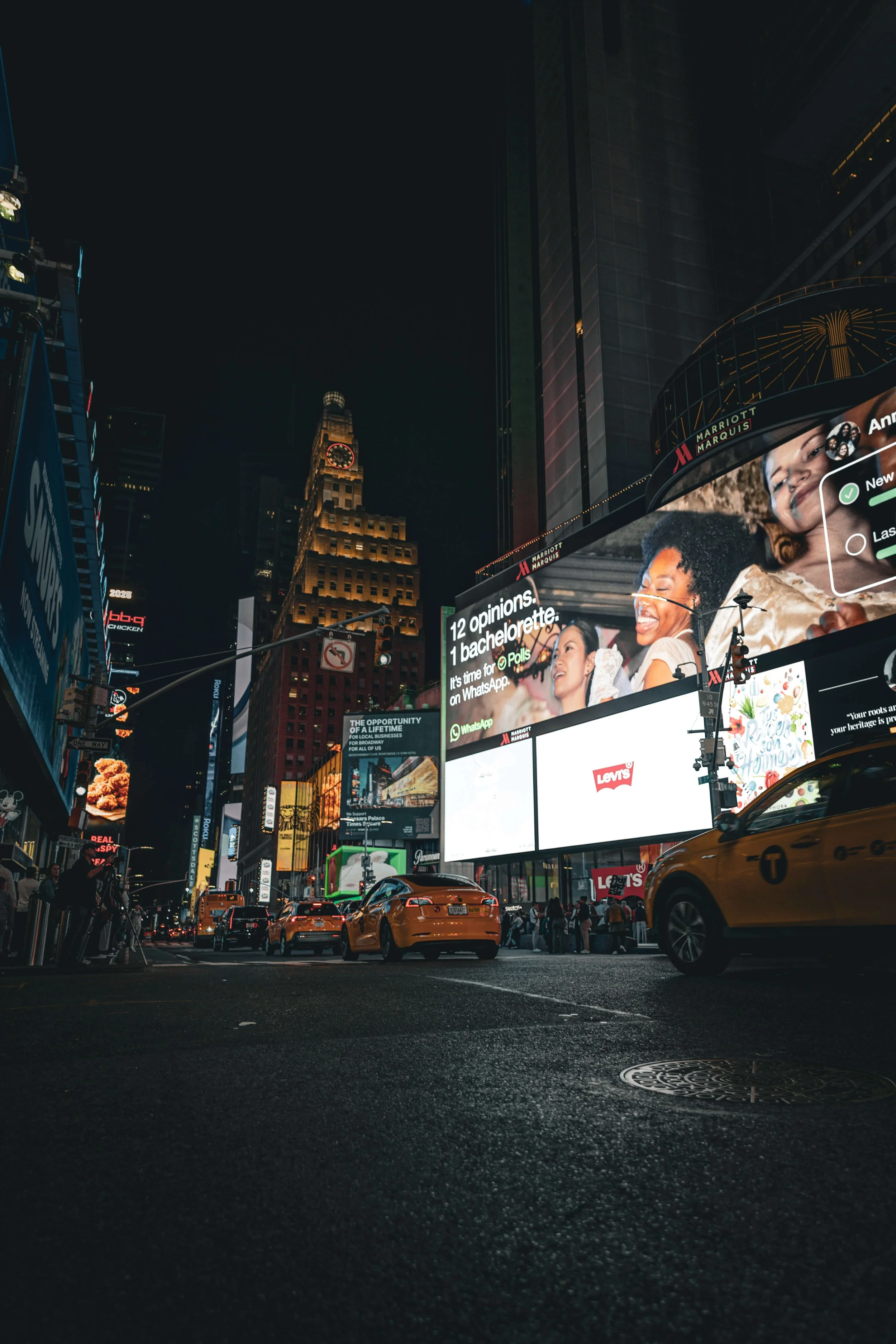 A nighttime city street scene with illuminated billboards, yellow taxis, cars, and pedestrians.