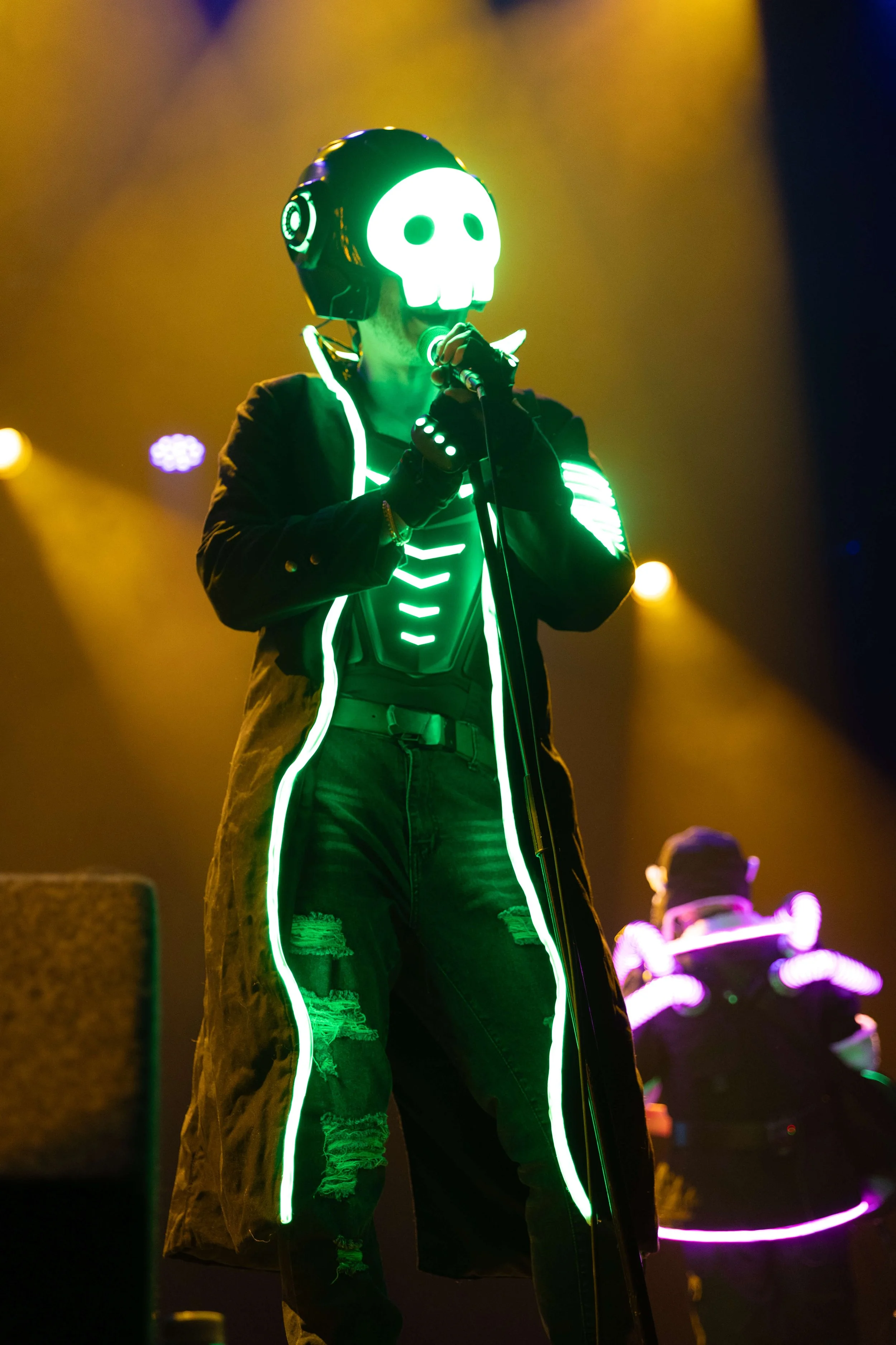 Person wearing a glowing skull mask, illuminated green, performing on stage with a microphone, in a black jacket with neon accents, jeans, and a long coat with neon outlines, in a dark setting with colorful lights.