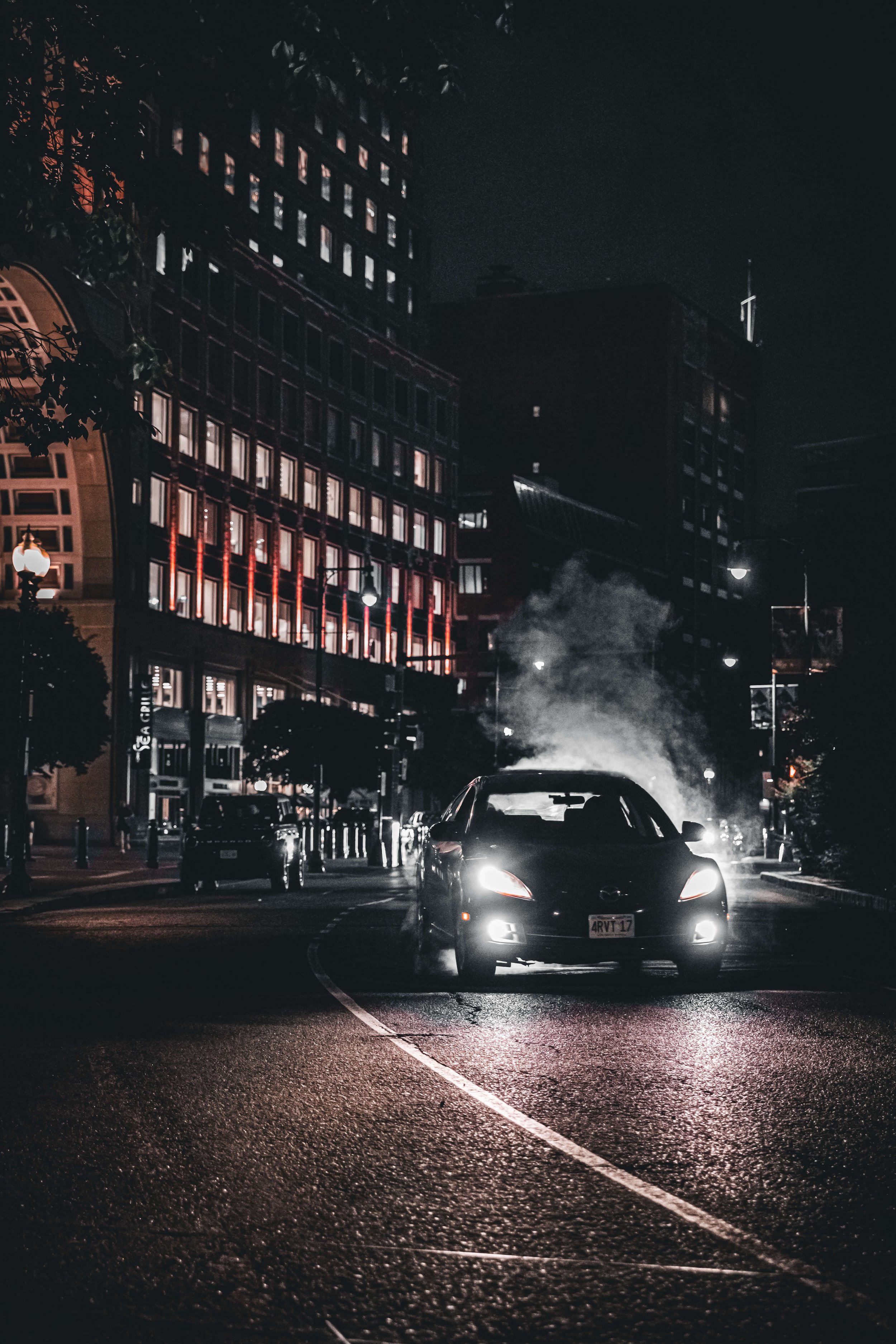 A black Mazda car driving on a city street at night, emitting exhaust fumes with illuminated buildings in the background.